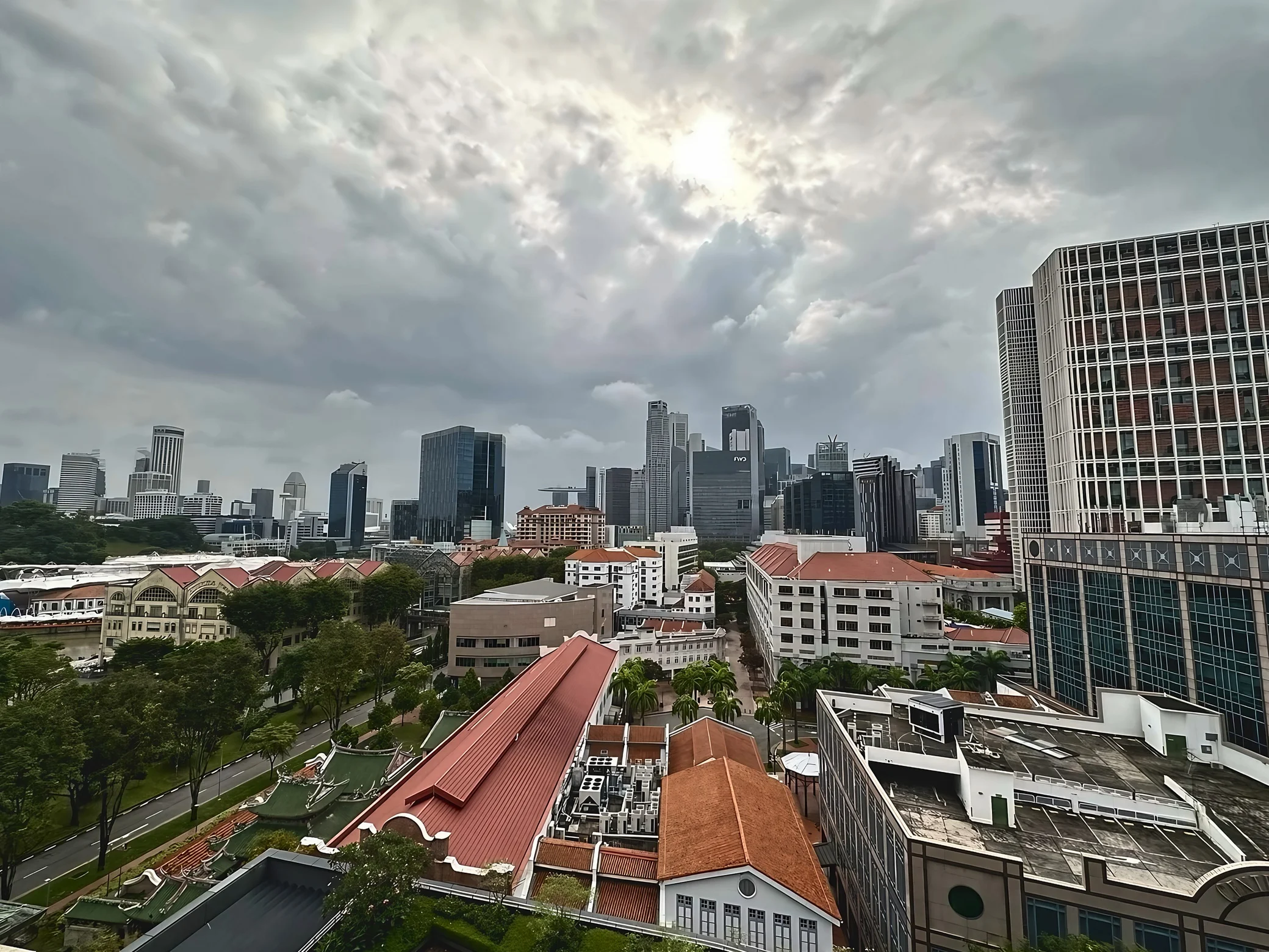 Red-roofed buildings, courtyards and trees fill the foreground beneath Singapore's skyline and a bright opening in heavy clouds.