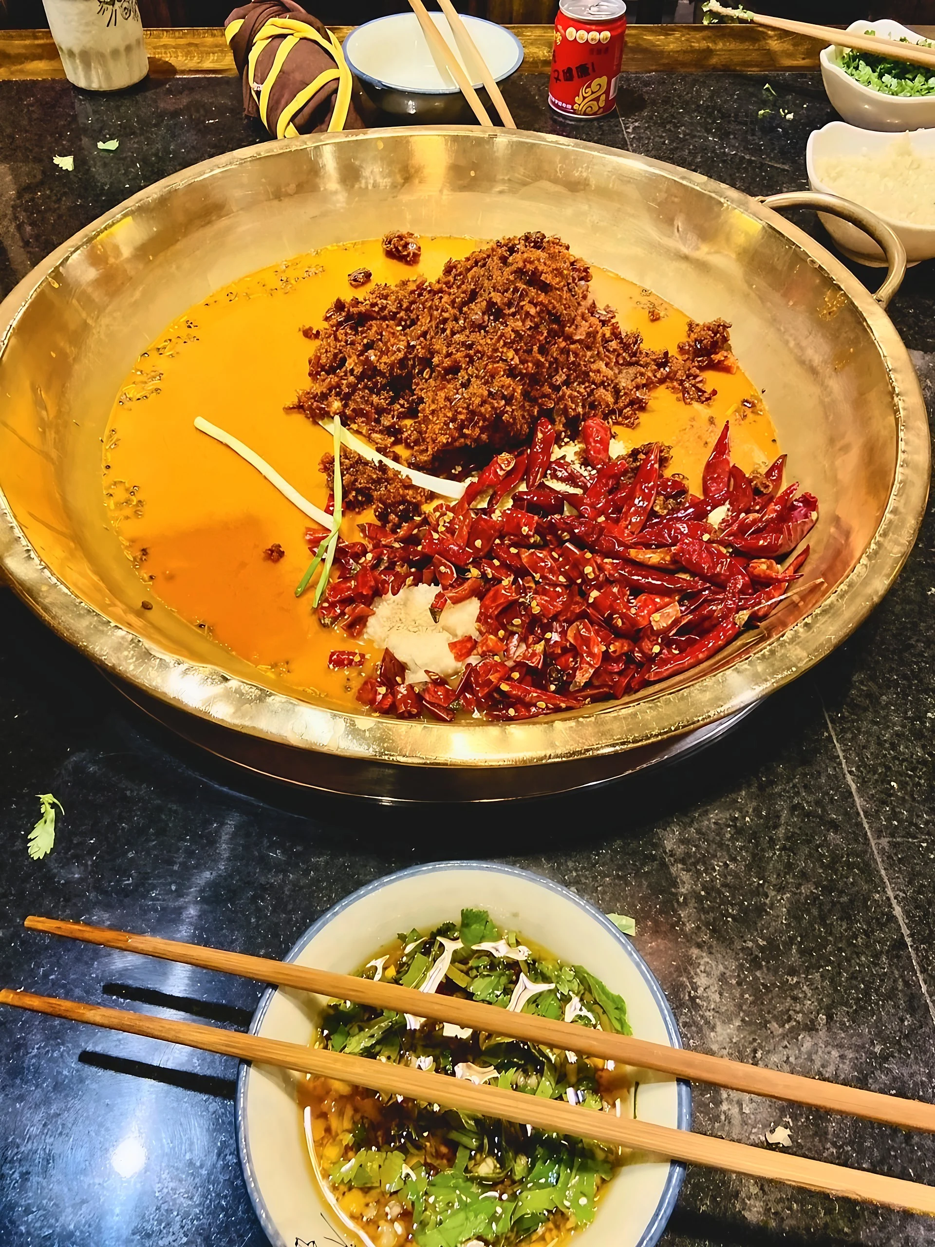 A copper hotpot in a Chengdu restaurant holds orange-red broth, dried chilies, minced garlic, scallions, and brown seasoning beside a cilantro dipping bowl.
