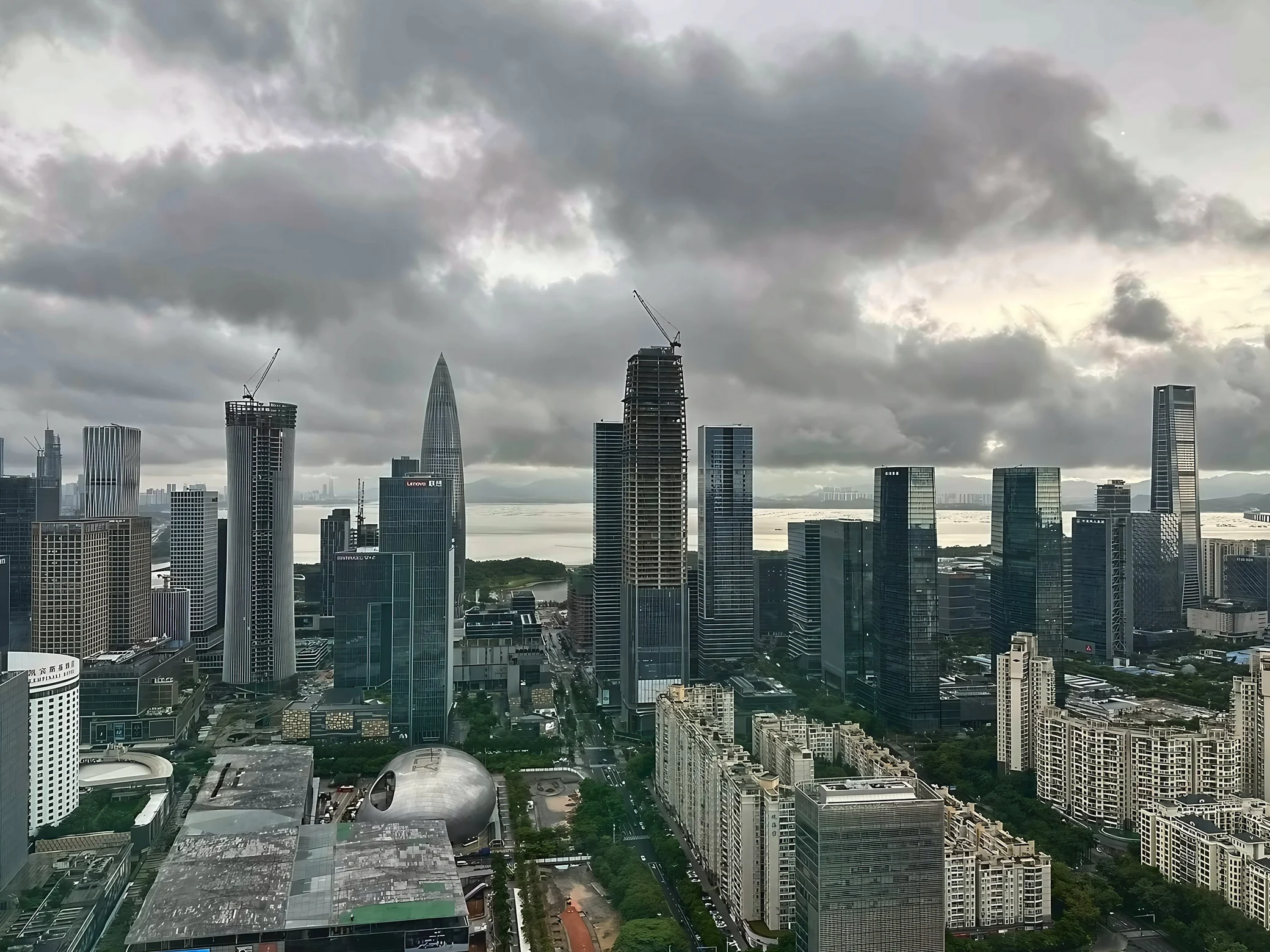 An elevated view of Shenzhen Bay shows China Resources Tower, surrounding skyscrapers and construction towers beneath heavy clouds, with the silver curved Shenzhen Poly Theatre in the foreground.