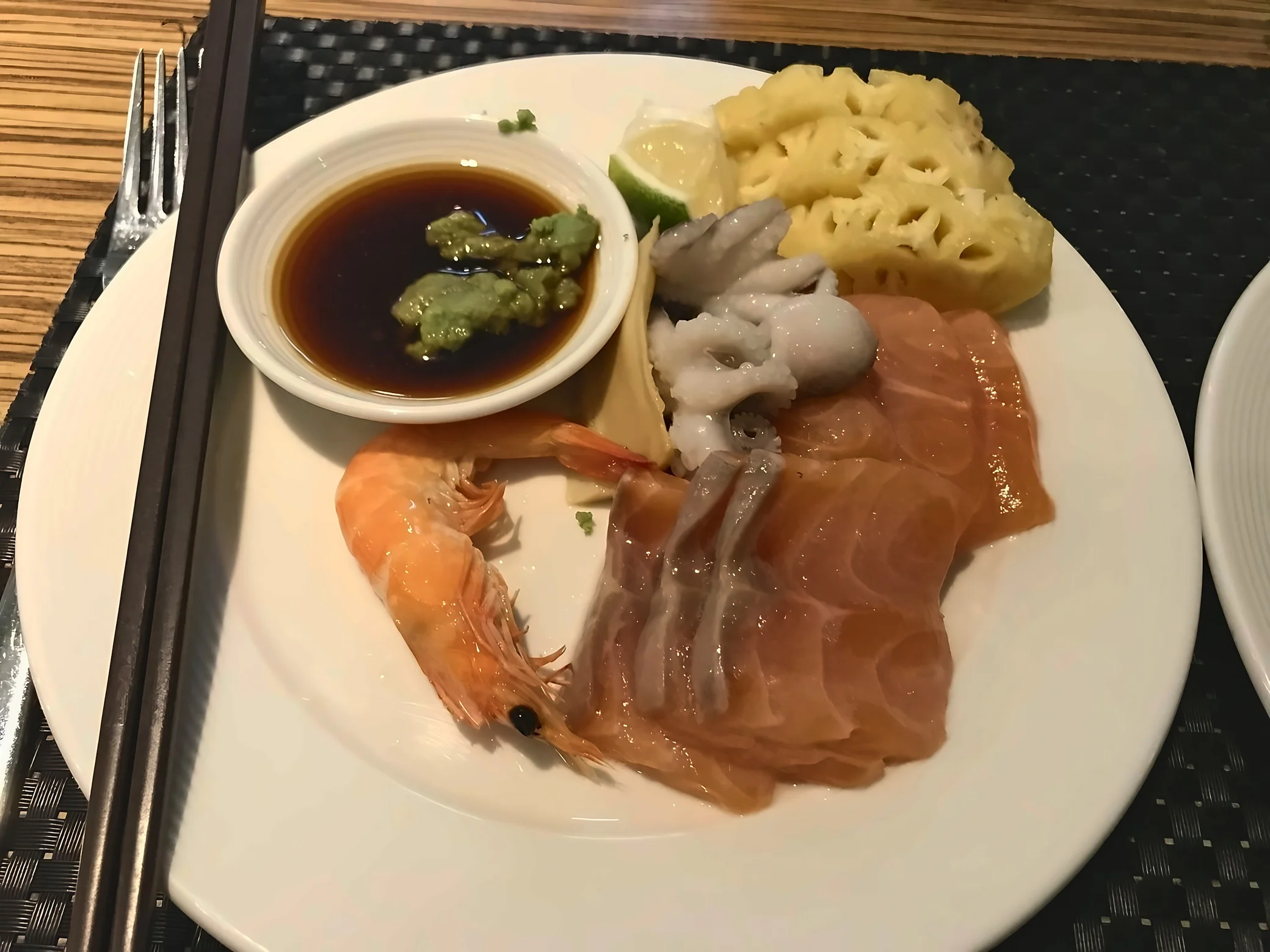 A white plate holds salmon sashimi, octopus, a whole shrimp and yellow garnishes beside soy sauce, wasabi and a citrus wedge.