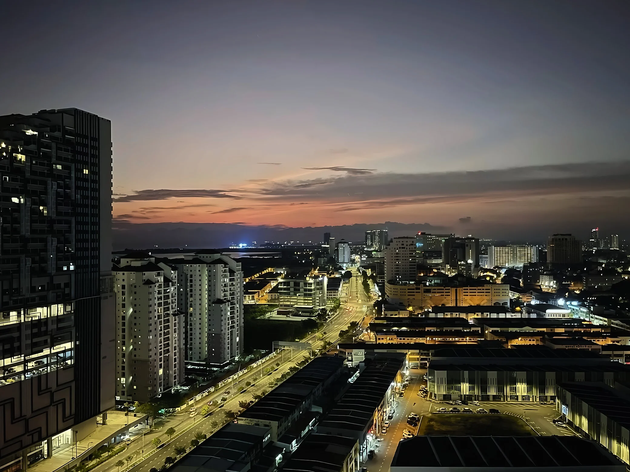 Illuminated roads and buildings in Malacca lead toward a distant strip of sea beneath orange clouds.