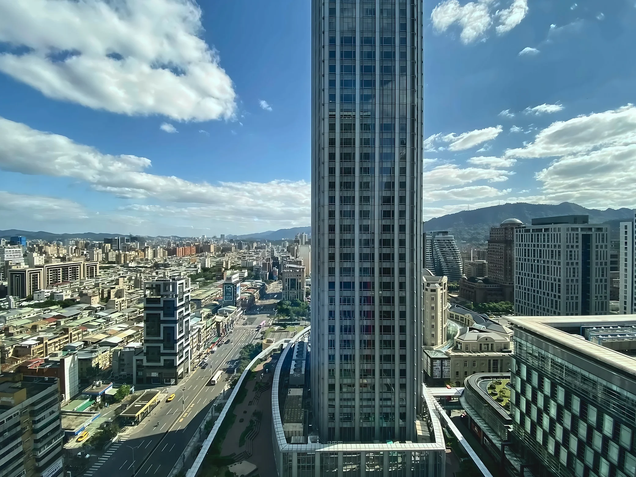 Taipei Nanshan Plaza overlooks city traffic and curved landscaped terraces, with distant hills beneath broad white clouds.