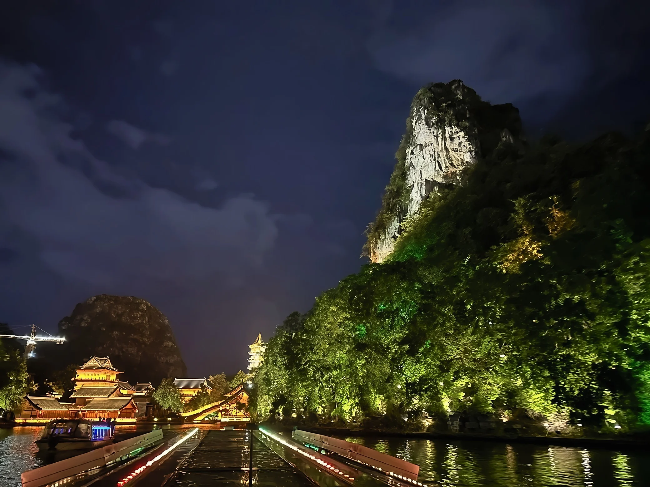 An illuminated karst peak, wooded shoreline, Chinese pavilions, a covered bridge, and a sightseeing boat frame the lake beyond two converging rows of boat lights.