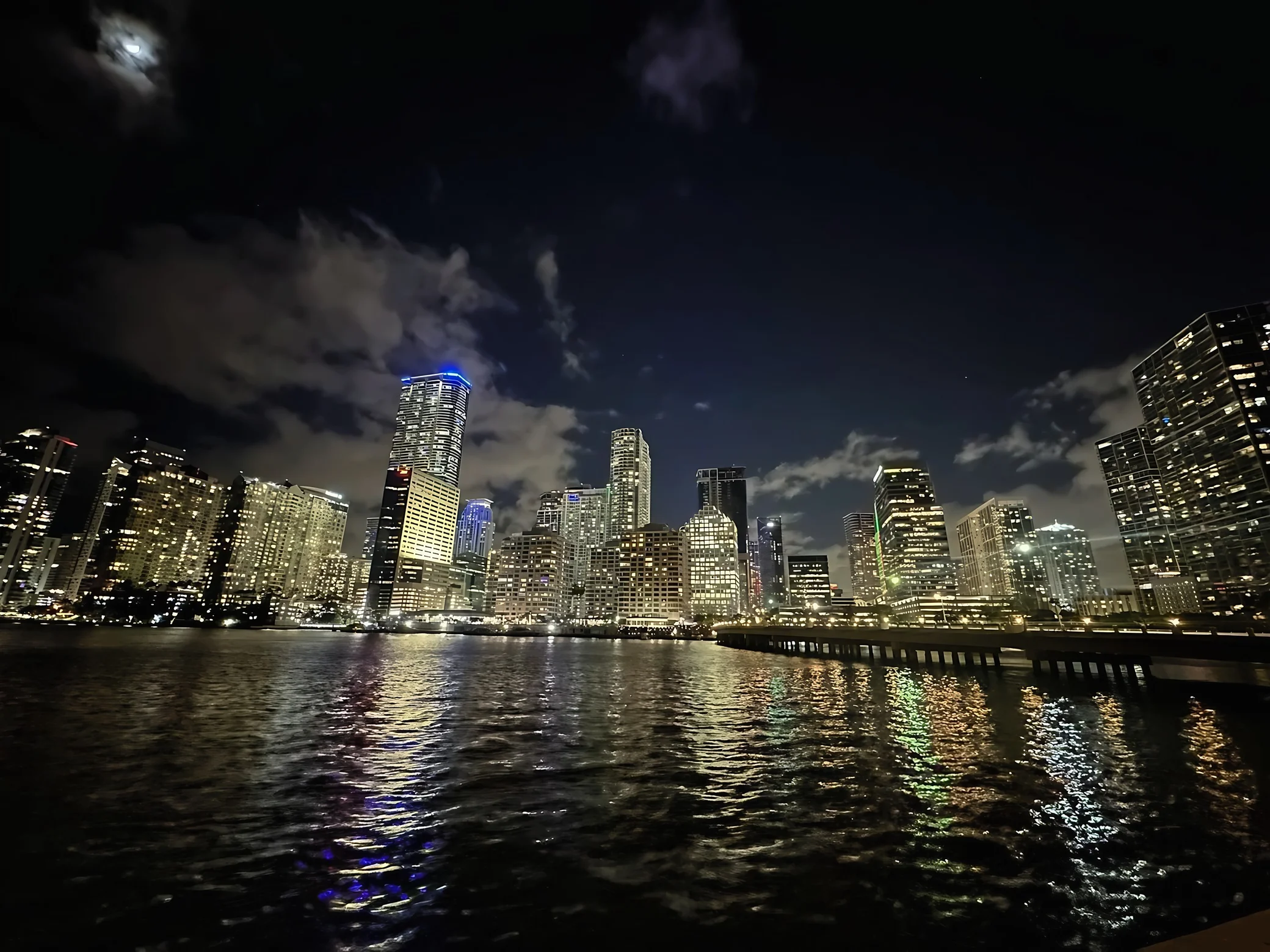 A moonlit waterfront skyline glows beneath scattered clouds, casting colorful reflections across the water beside a pier.