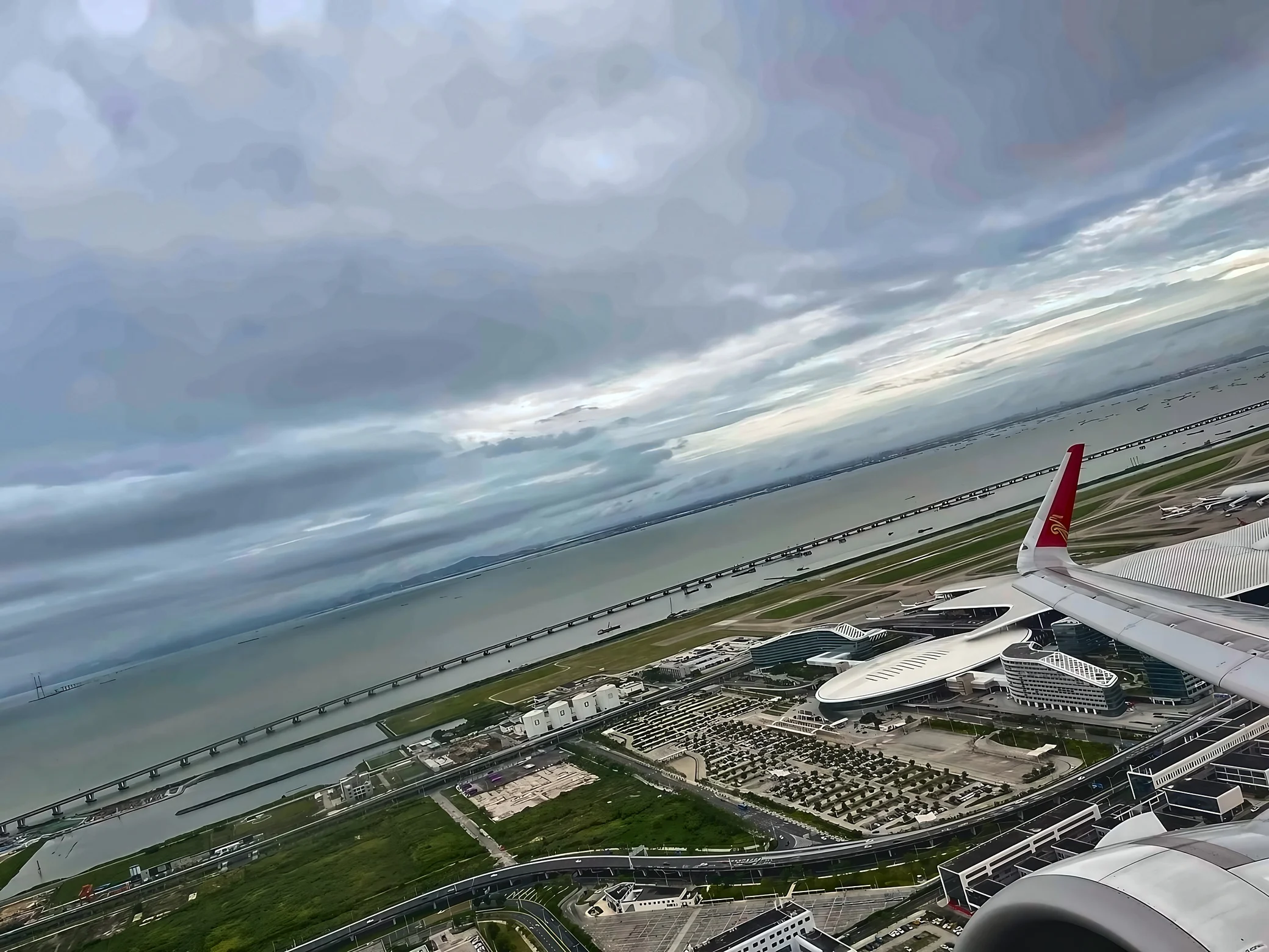 An airplane wing and part of its engine frame an aerial view of Shenzhen Bao'an International Airport, coastal roads, bridges, and the sea.