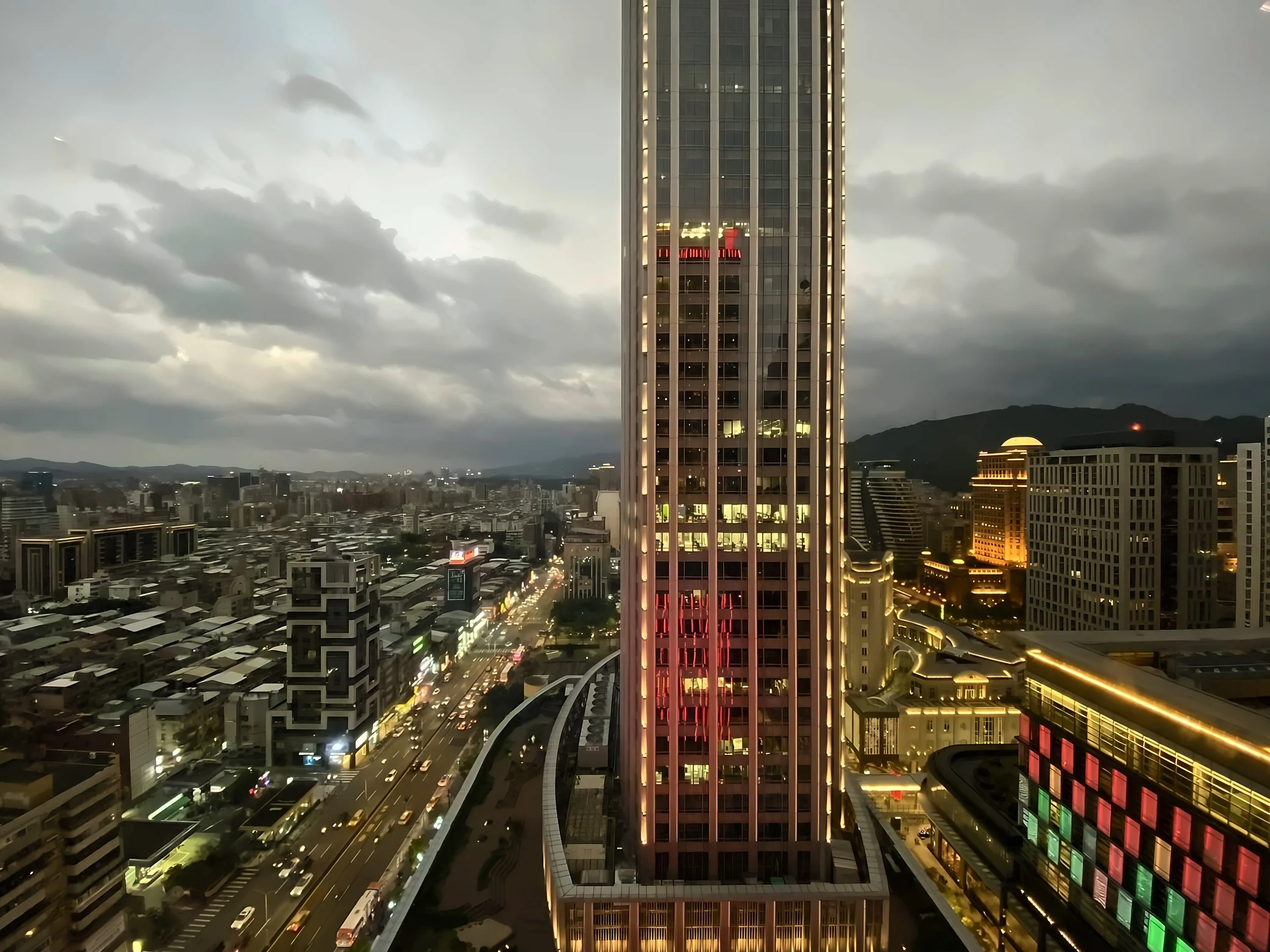 An illuminated Taipei tower rises beside busy streets, dense city blocks, colorful mall lighting, and distant hills under layered clouds.