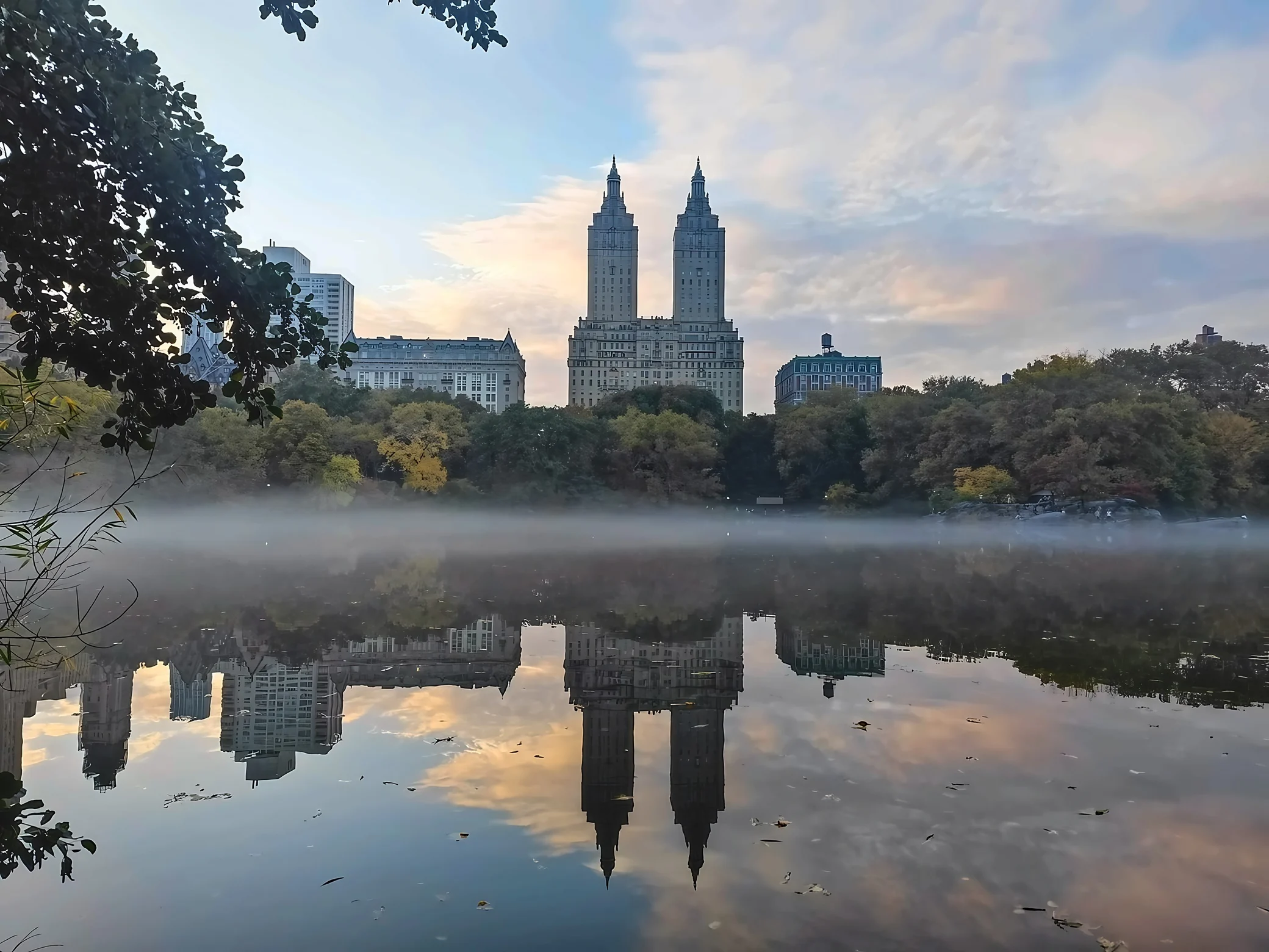 The San Remo towers stand near the center above a misty Central Park lake reflecting autumn trees and pastel clouds.