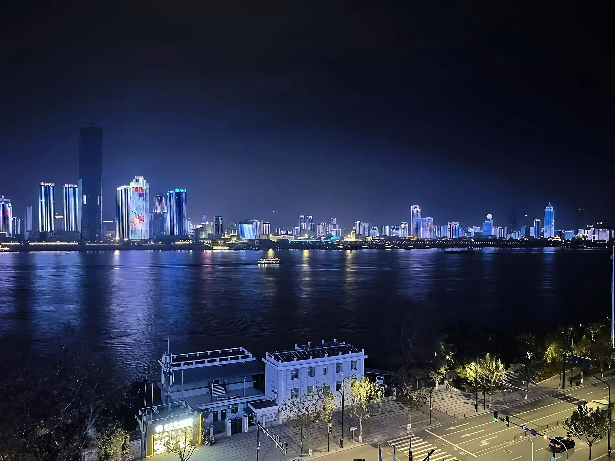 Blue and white skyline lights reflect across the Yangtze River around a lit cruise boat, with low-rise buildings, trees and a road along the near bank.
