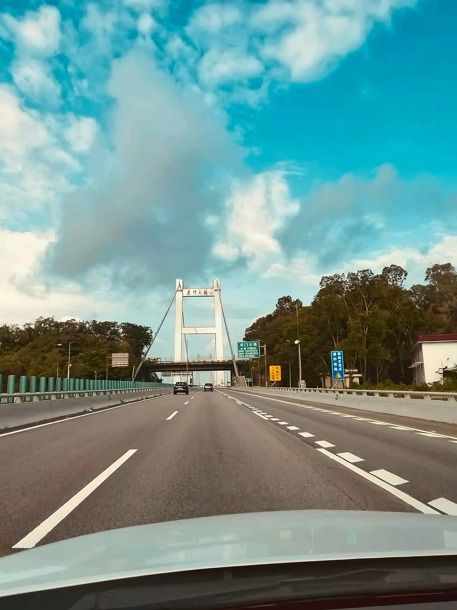 The white tower and suspension cables of Humen Bridge rise ahead of two dark vehicles beneath a dramatic cloud-filled sky.