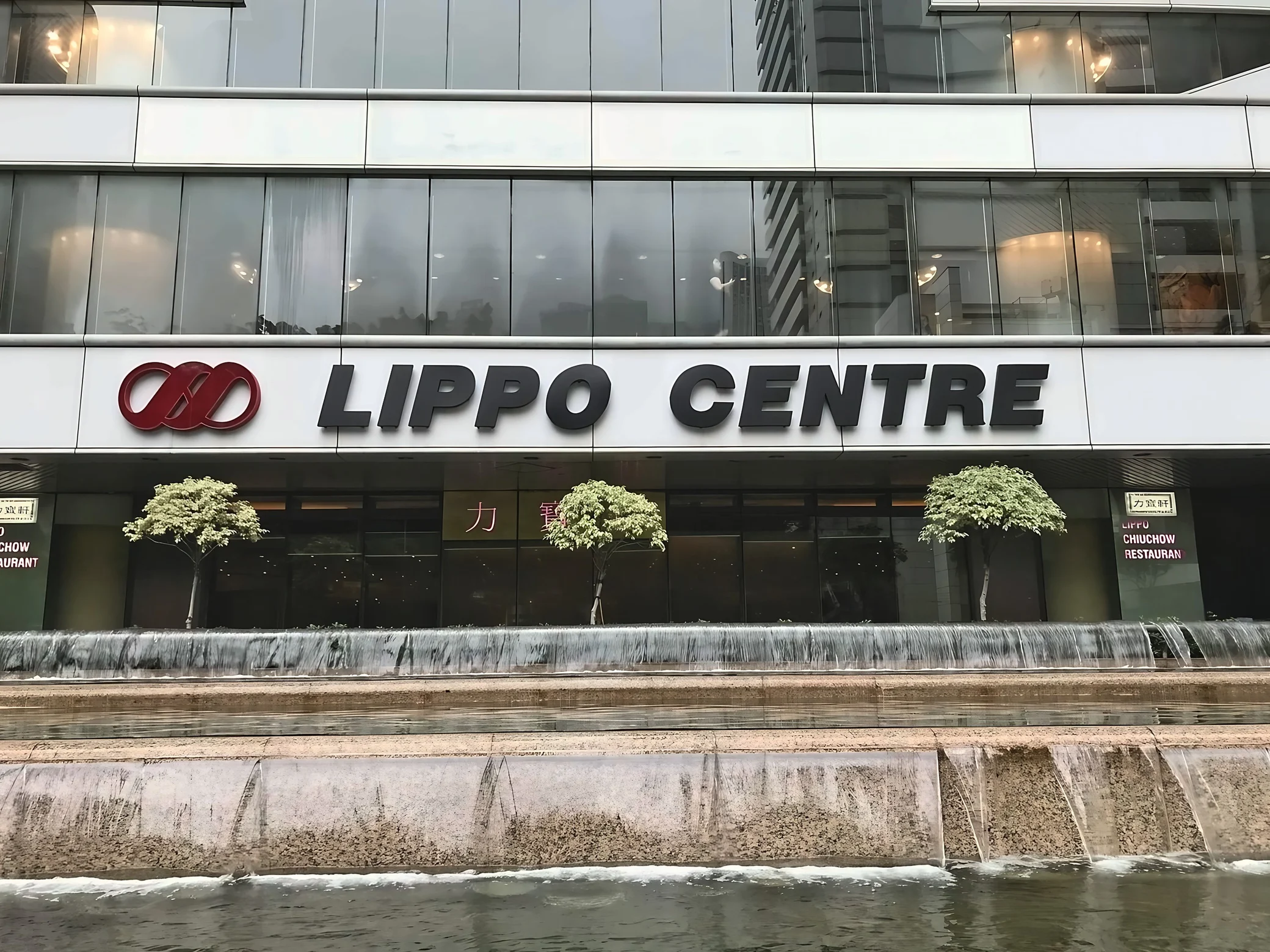 The Lippo Centre facade, with its large lettering and red logo, stands behind trimmed trees and a tiered water feature.