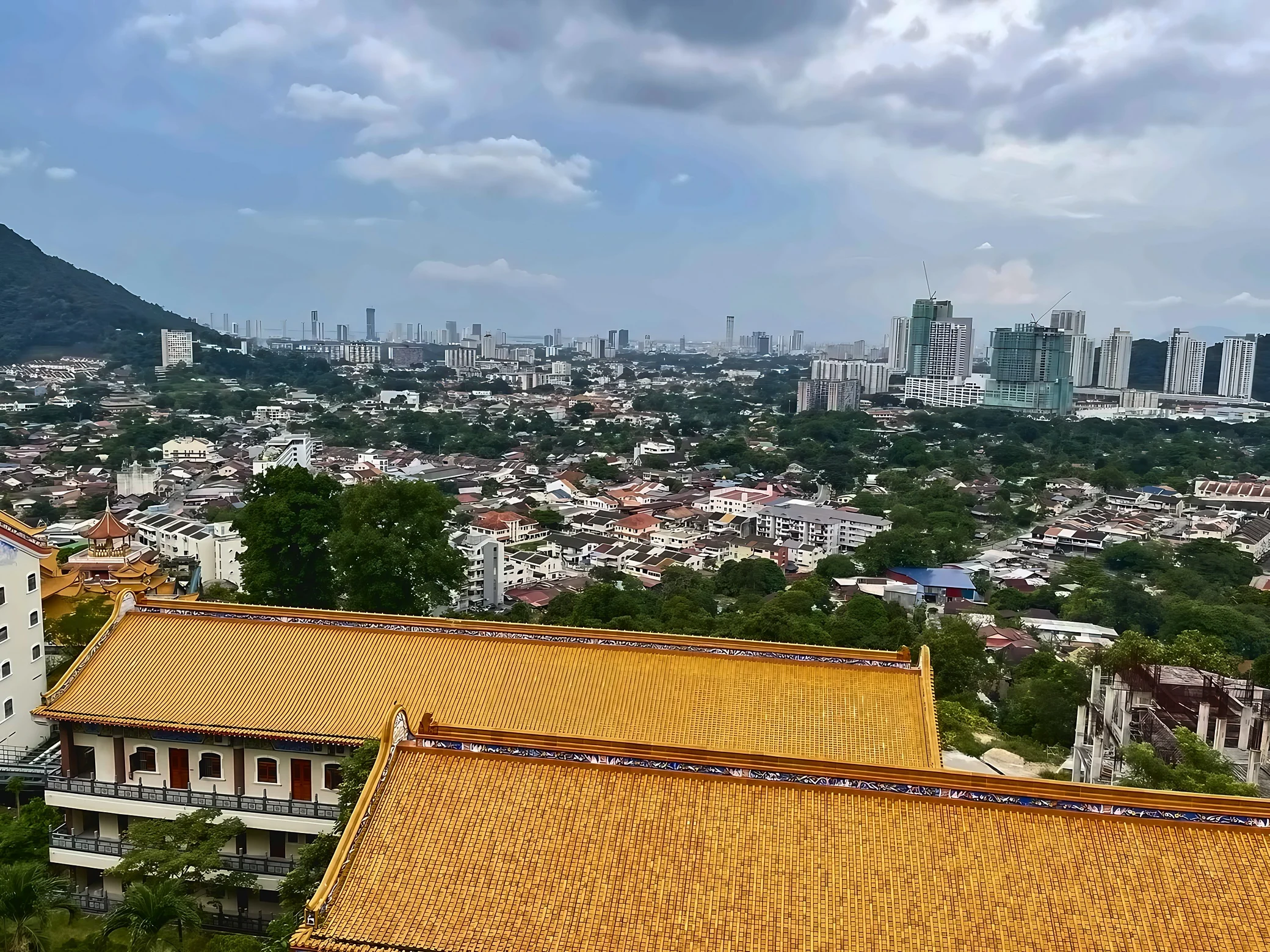 Expansive golden roofs at Kek Lok Si overlook leafy residential neighborhoods, distant high-rises and a green hillside beneath a cloudy sky.