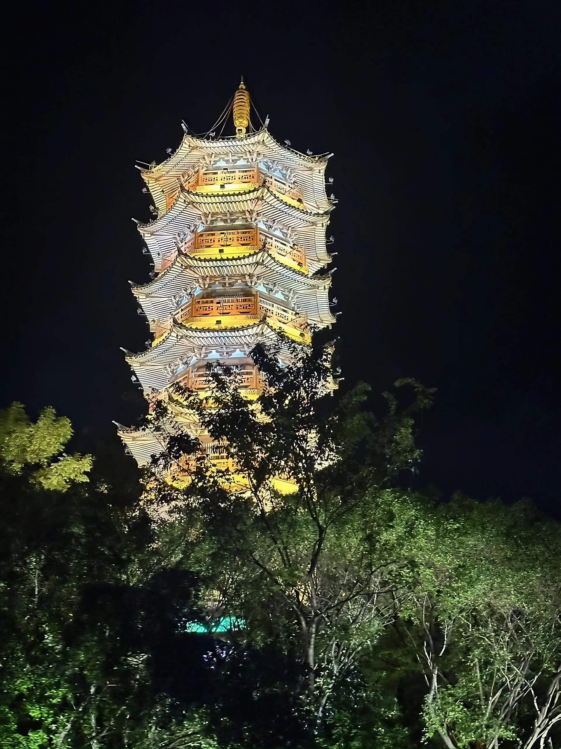 A multi-tiered pagoda glows with white and warm yellow lighting above illuminated trees that partly obscure its lower levels.