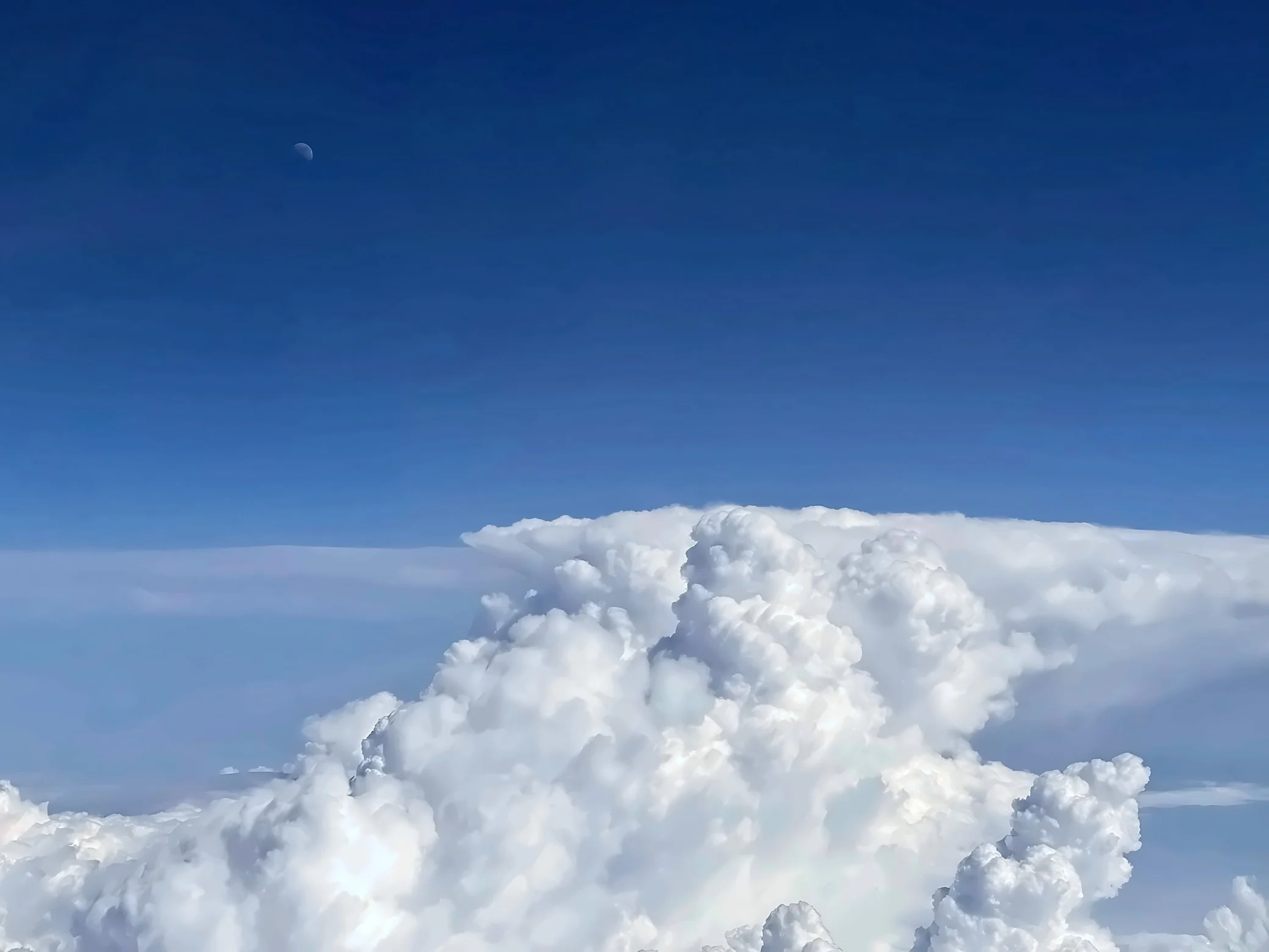 Towering white clouds spread into an anvil shape beneath a deep blue sky with a faint moon in the upper left.
