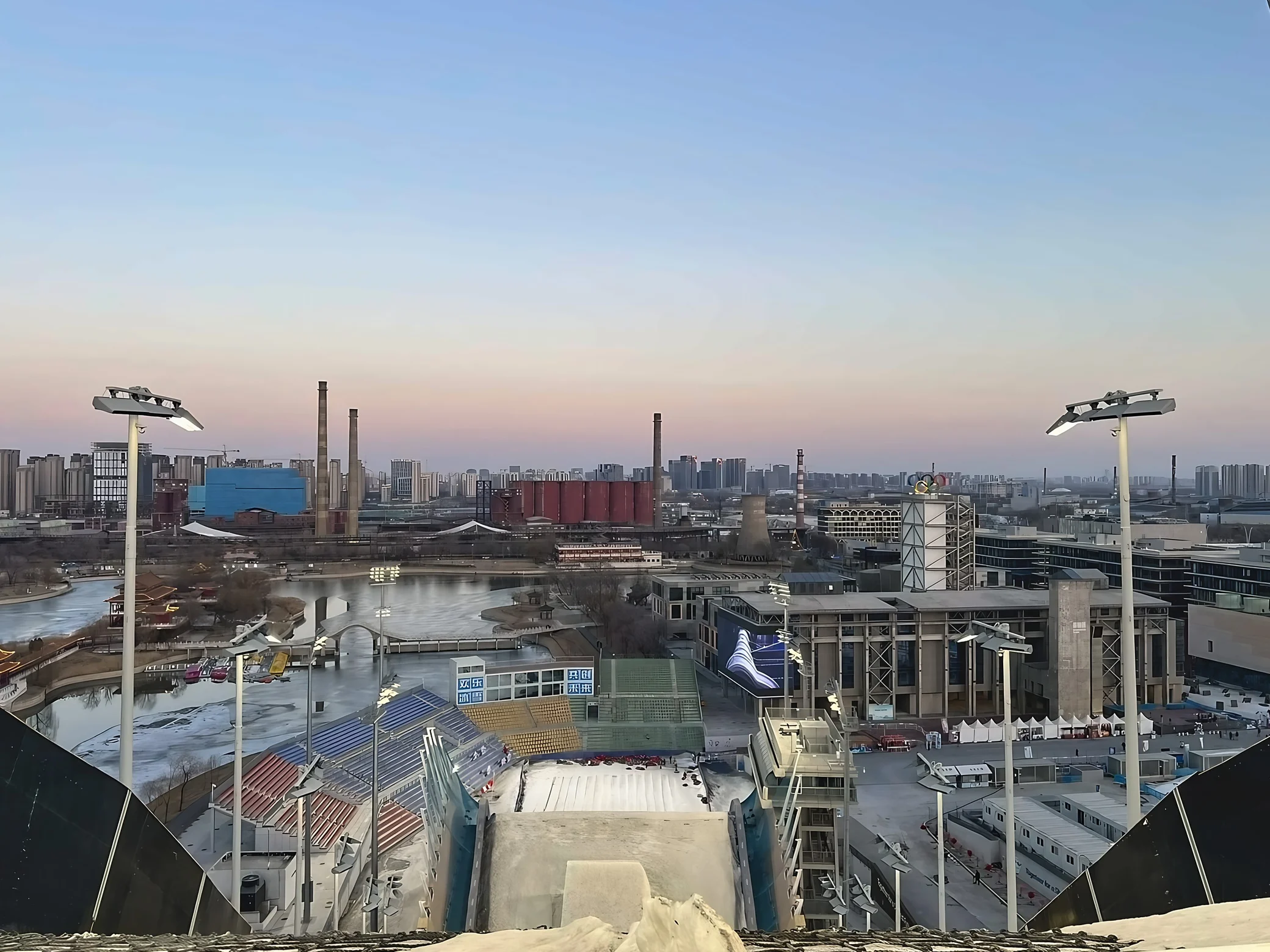 The snowy slope of Shougang Big Air overlooks colorful stands, a frozen lake, industrial chimneys and a city skyline beneath a pale blue and pink sky.