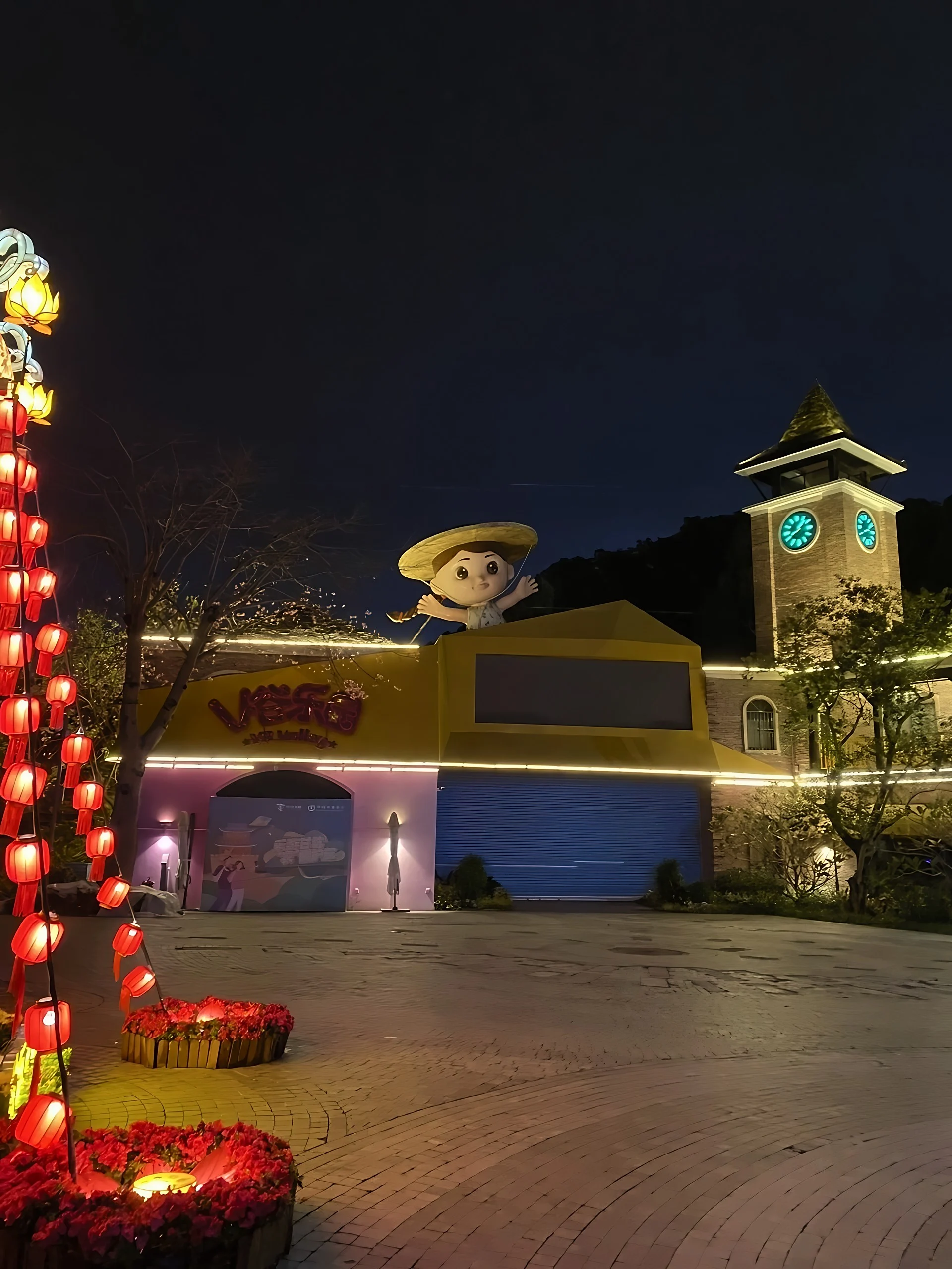 A straw-hatted cartoon figure tops a yellow-and-pink building beside a glowing clock tower and strings of red lanterns in Gankeng Ancient Town.