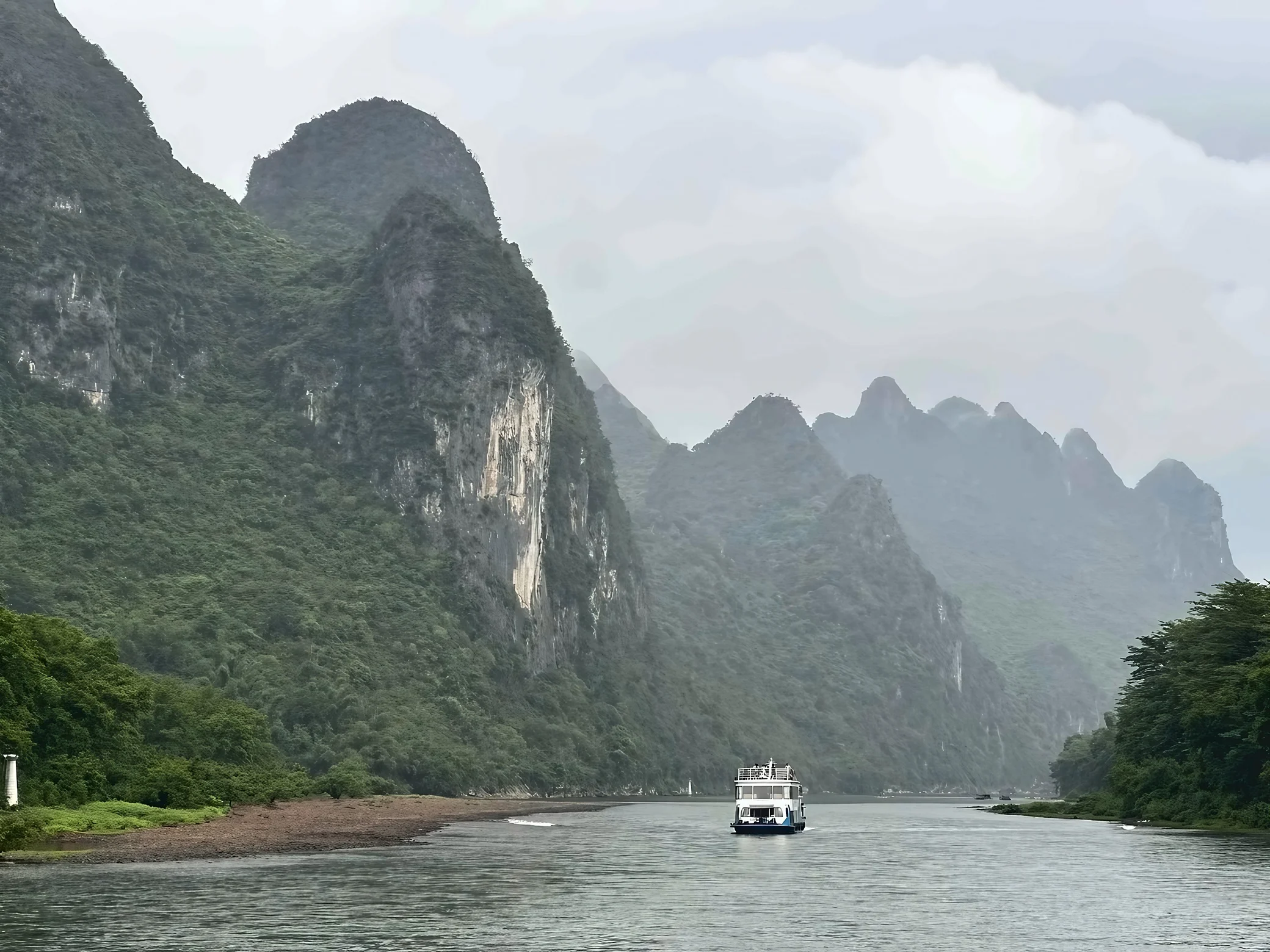 A white tour boat travels between steep limestone peaks along a hazy river bordered by trees and a gravel bank.
