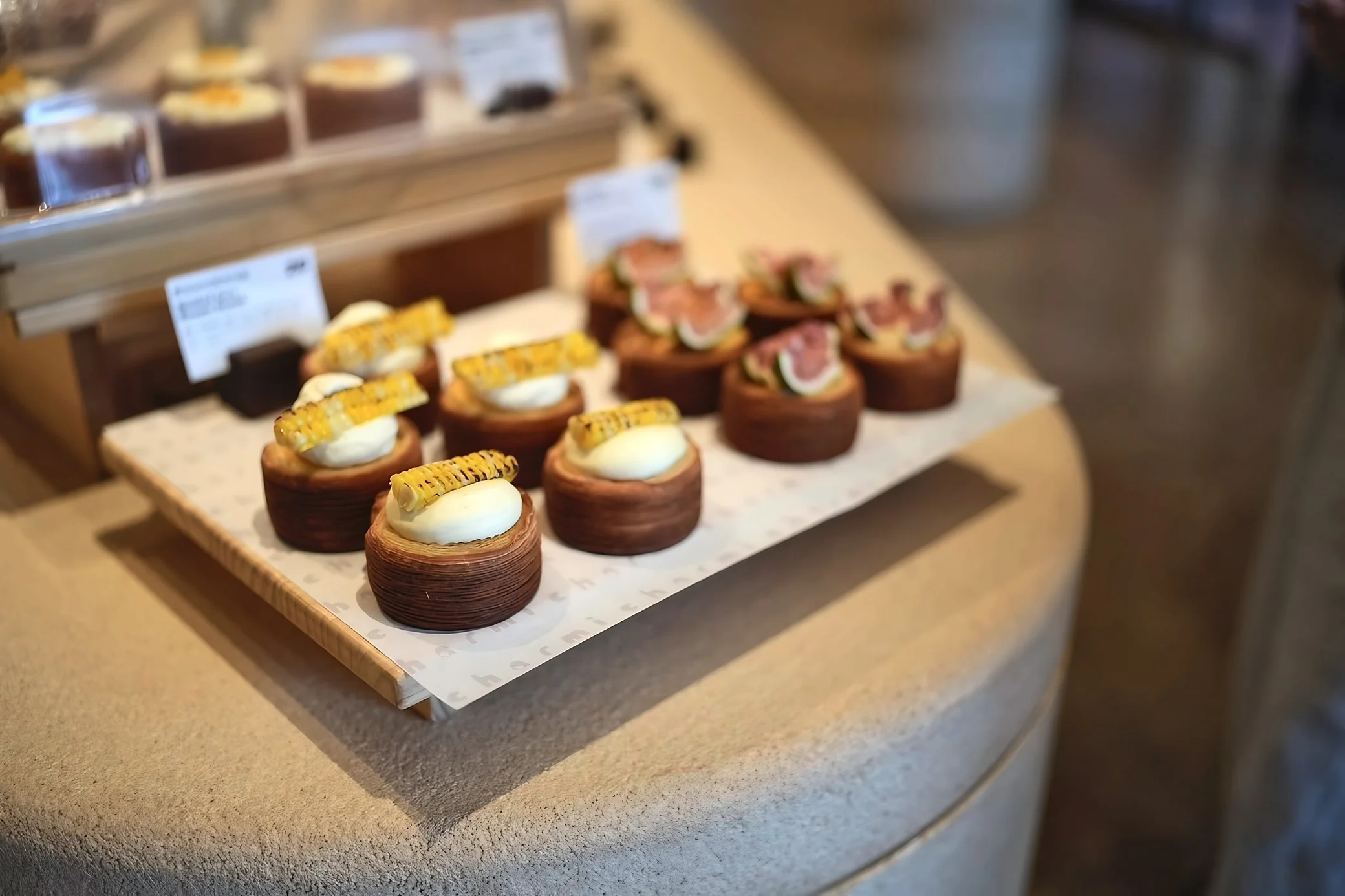 Cylindrical layered pastries topped with cream and charred corn or sliced figs sit on a tray against a softly blurred bakery display.