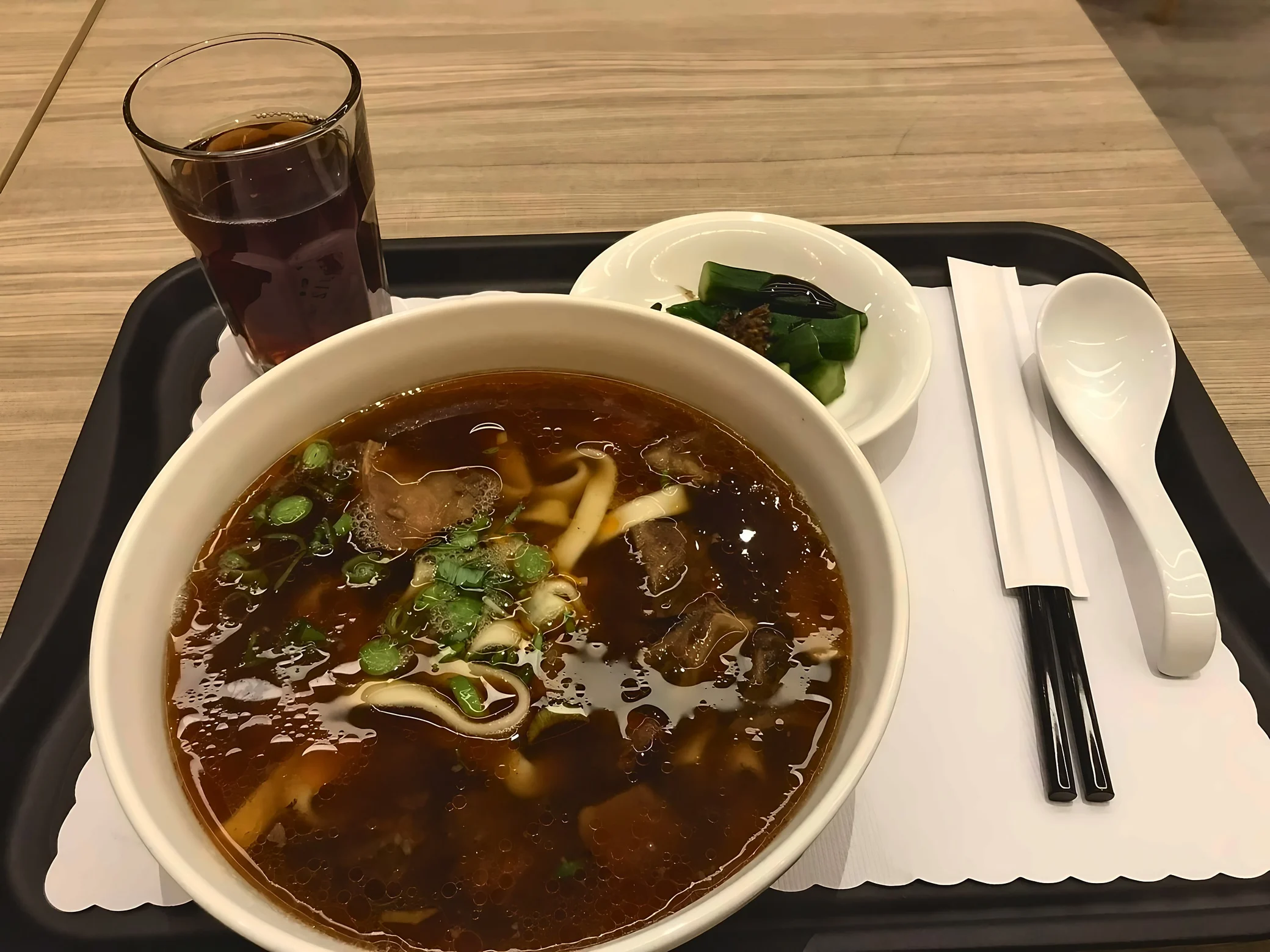 A tray holds beef noodle soup, a cucumber side dish, a dark drink, chopsticks, and a white soup spoon.