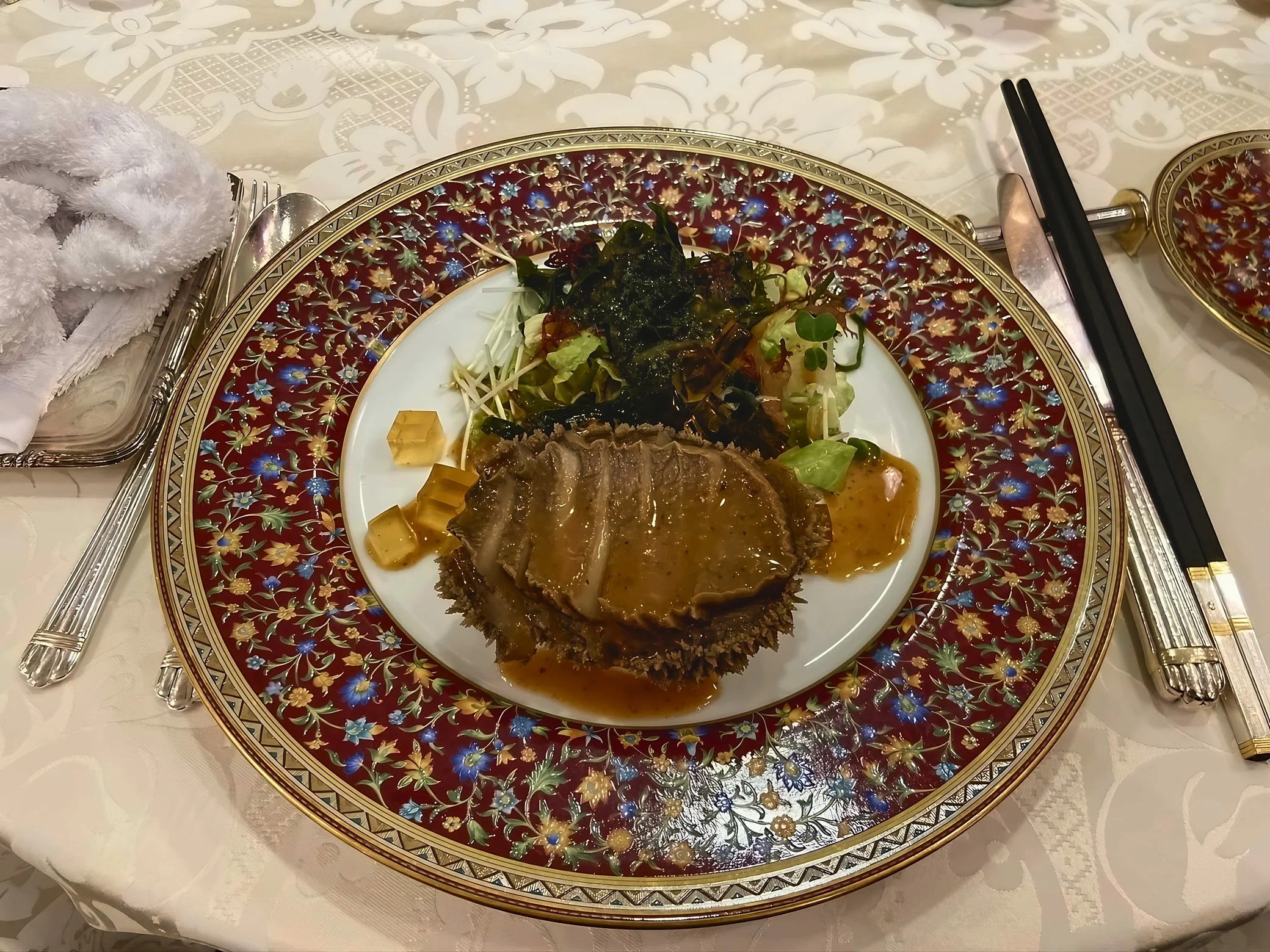 A close-up shows sauced abalone with seaweed and leafy salad on a richly patterned floral plate.
