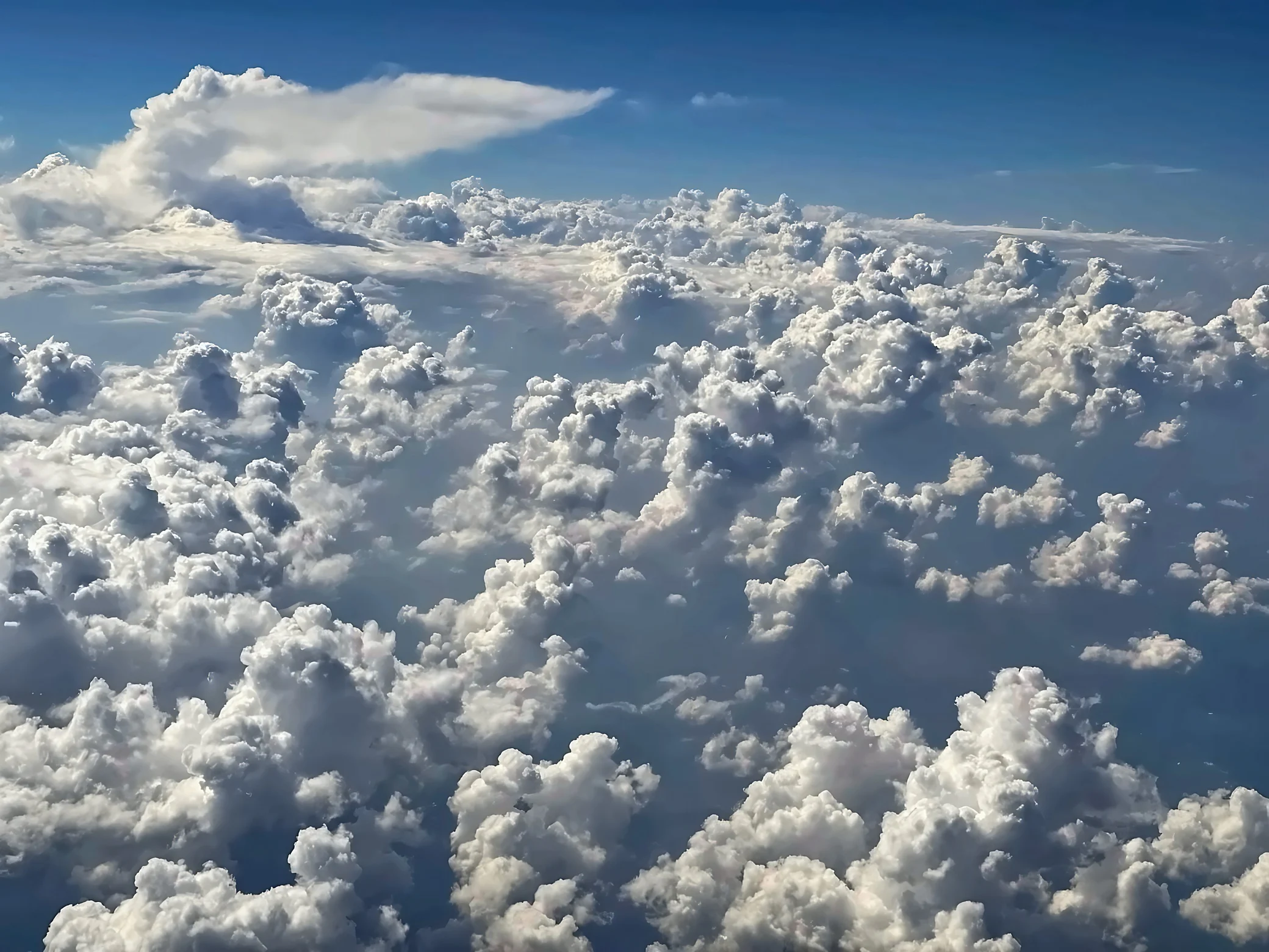 Layered white cumulus clouds fill the view beneath a blue sky, with an anvil-shaped cloud rising in the distance.