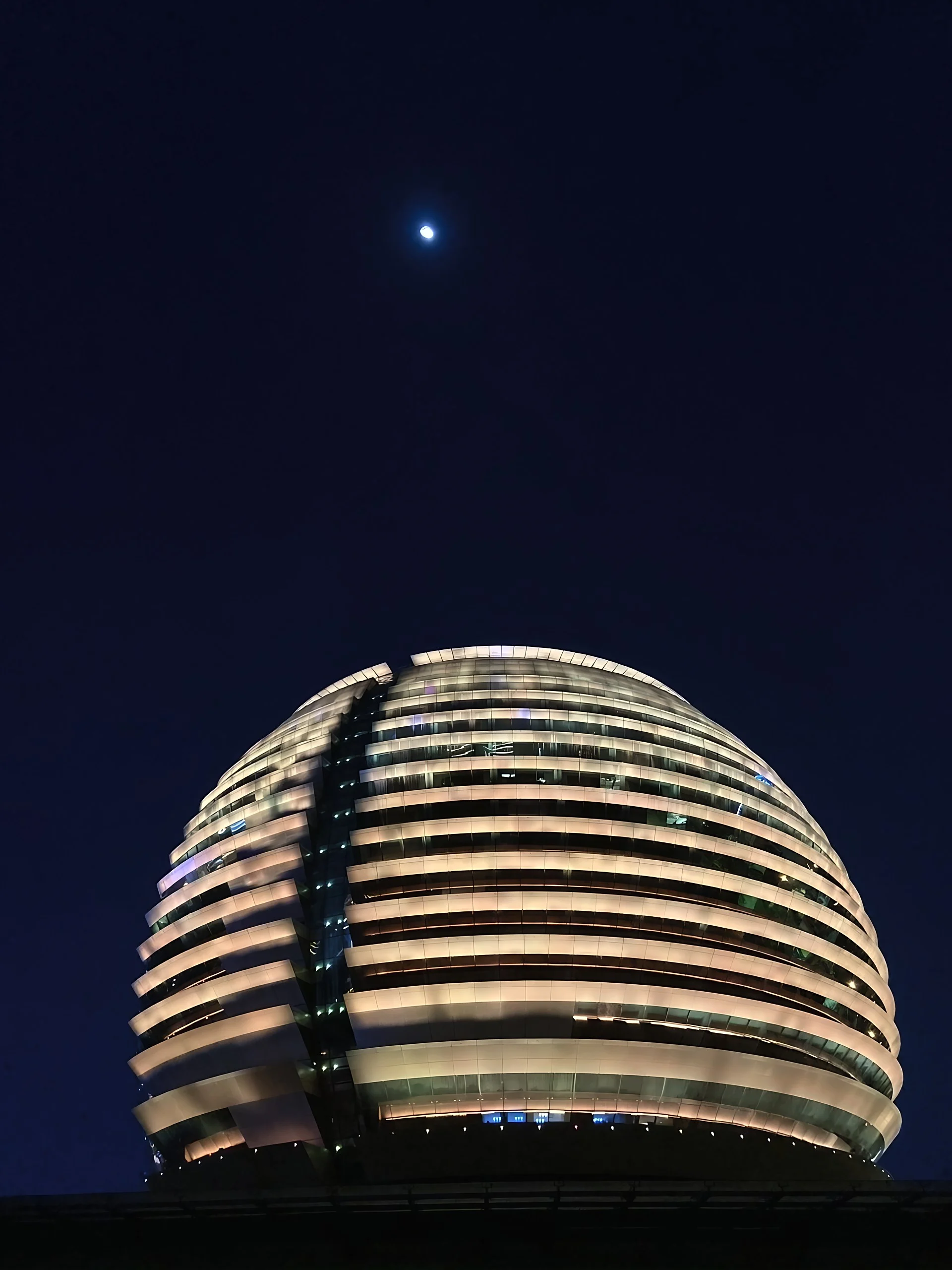 A bright moon hangs above the illuminated spherical facade of Hangzhou International Conference Center against a dark blue sky.