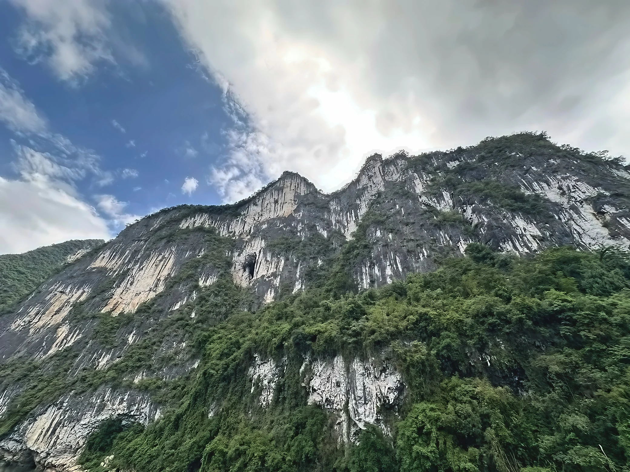 A towering limestone cliff covered in greenery rises beneath blue sky and broad clouds.