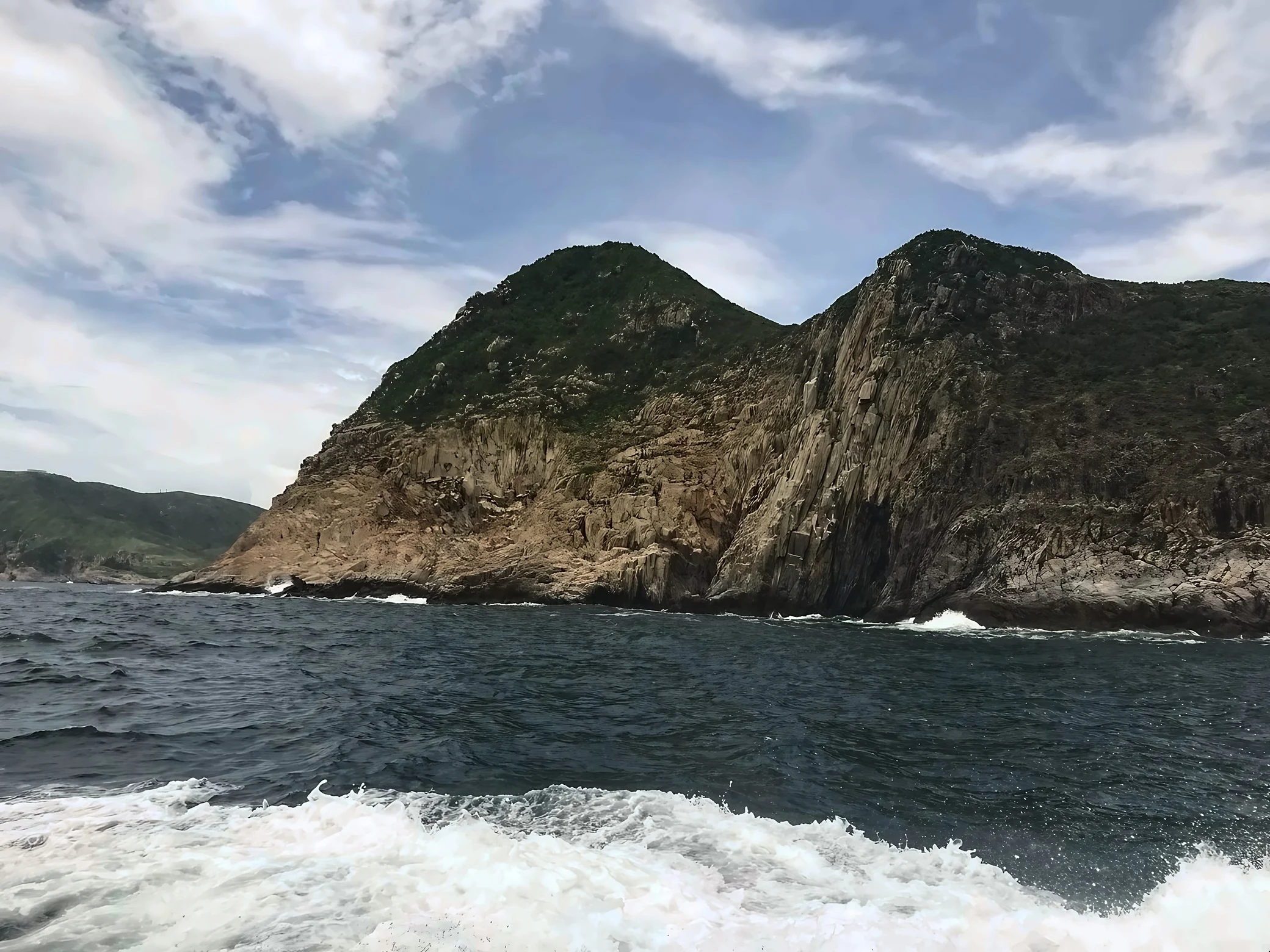 Green hills and vertically textured coastal cliffs overlook dark seawater, with small waves at the rocks and a broad band of foamy boat wake in the foreground.