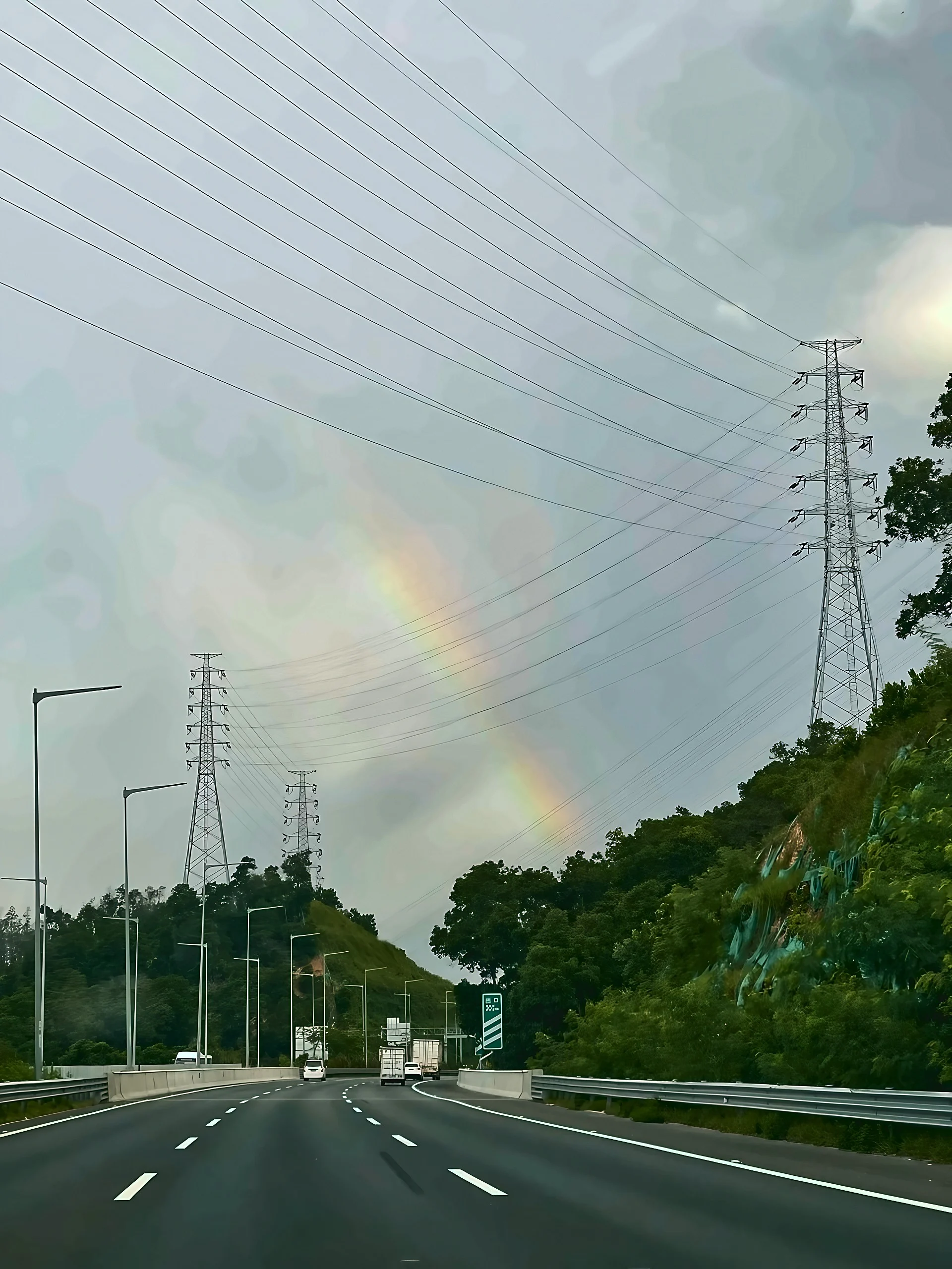 A rainbow overlaps power lines above a multilane highway running between green slopes.