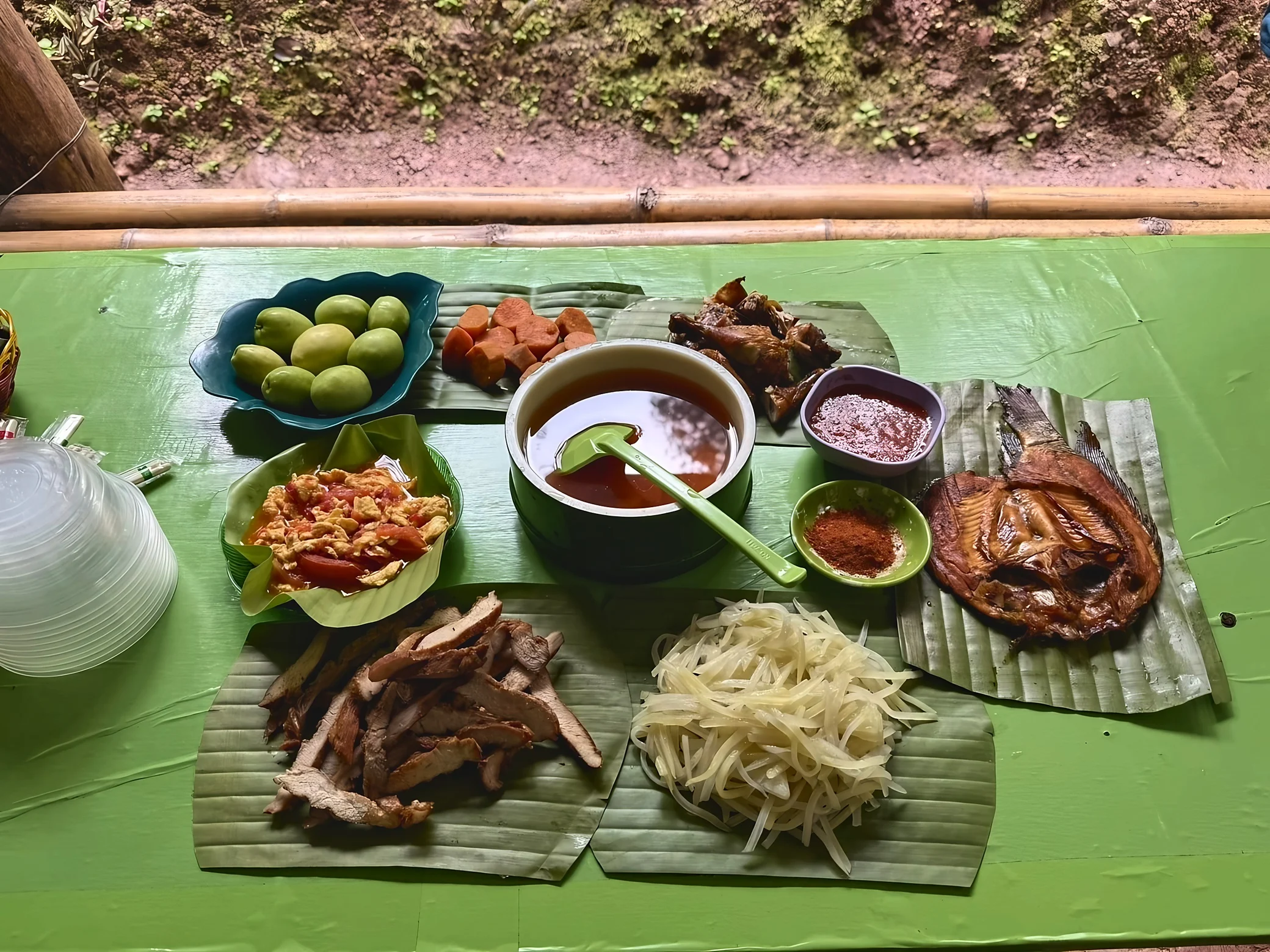 Fish, sliced meat, shredded vegetables, tomato and eggs, soup, fruit, and dipping sauces are arranged on banana leaves across a green table.