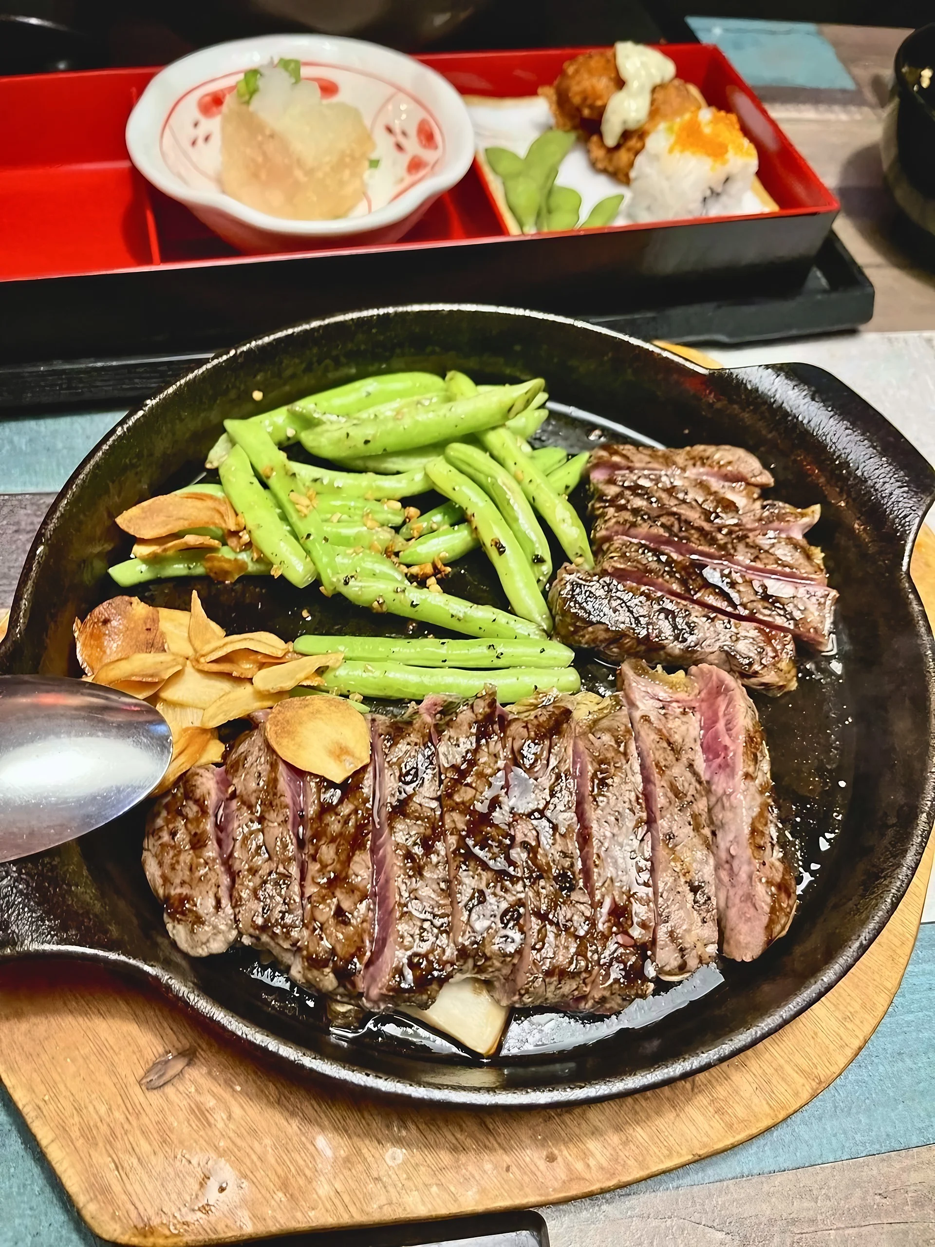 Sliced steak, green pea pods, and garlic chips fill a sizzling plate at a Penang restaurant, with sushi and fried side dishes behind it.