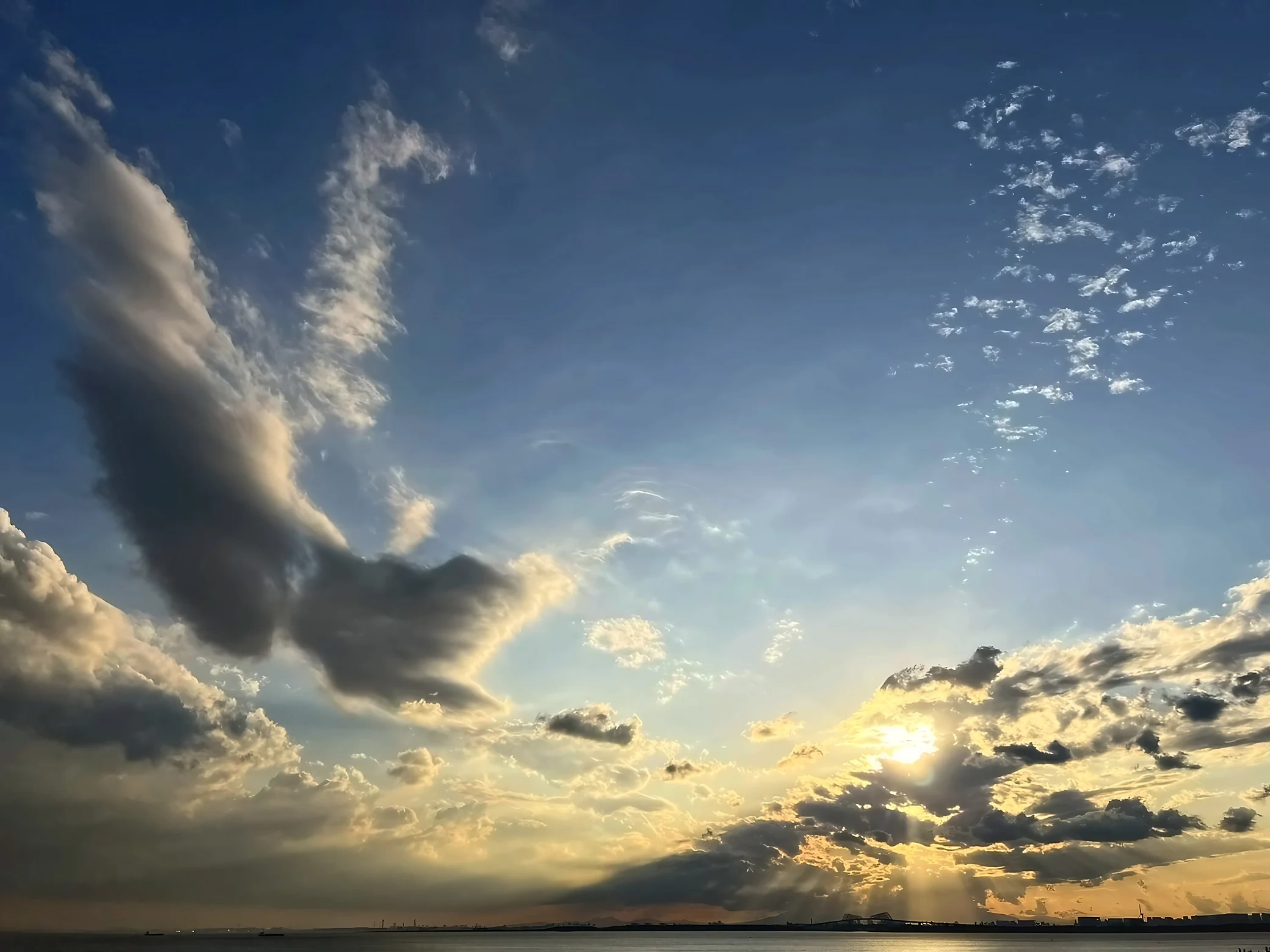 Dark clouds and golden cloud edges cross the blue sky while sunlight streams through toward the distant shoreline.