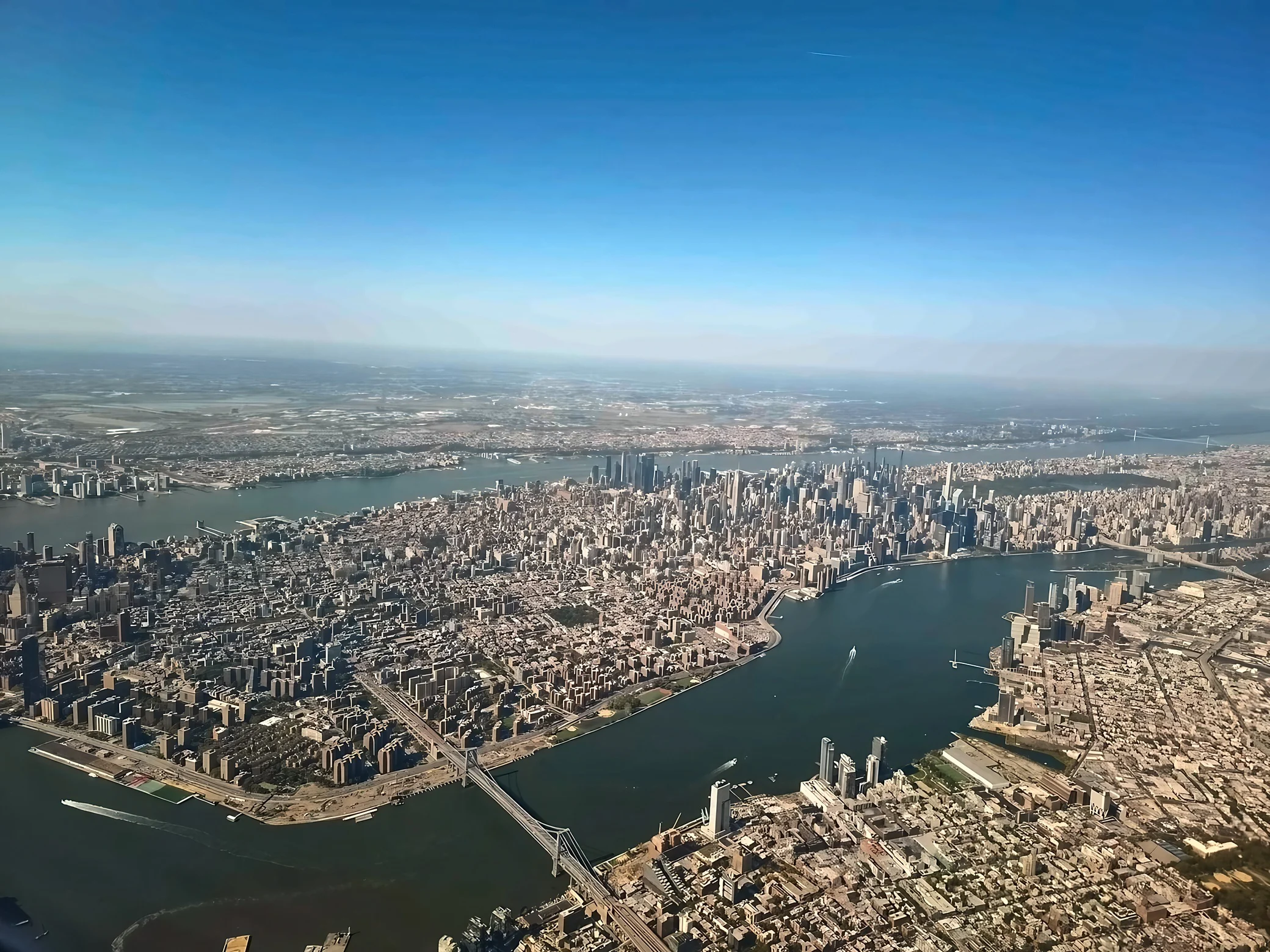 Manhattan's skyline stretches along the East River, with the Williamsburg Bridge crossing the foreground and boats leaving white wakes below.