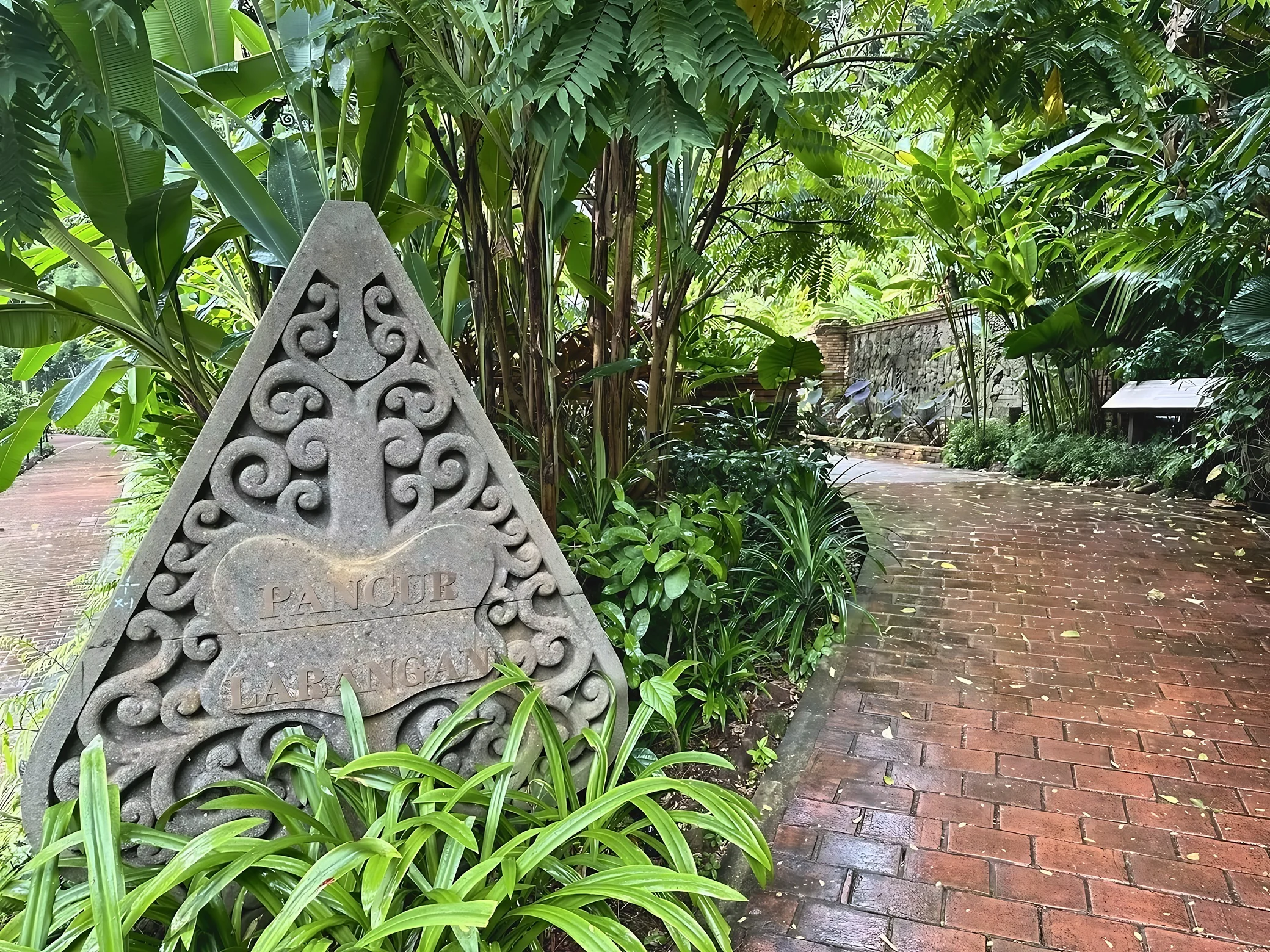 A carved Pancur Larangan stone marker stands among tropical plants beside a wet brick path in Fort Canning Park.