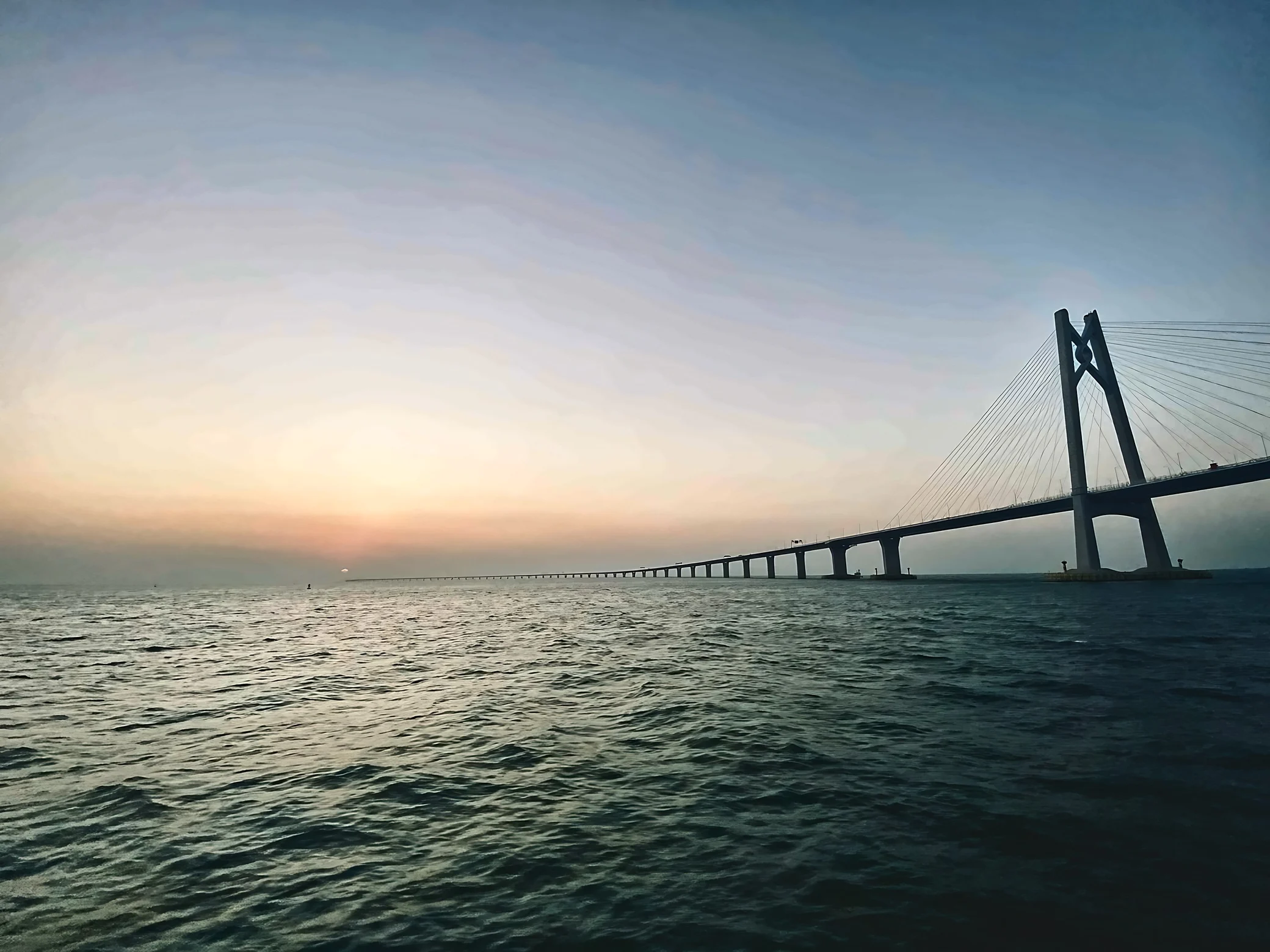 A cable-stayed sea bridge extends from a tall tower on the right toward a small red sun near the horizon above rippling water.