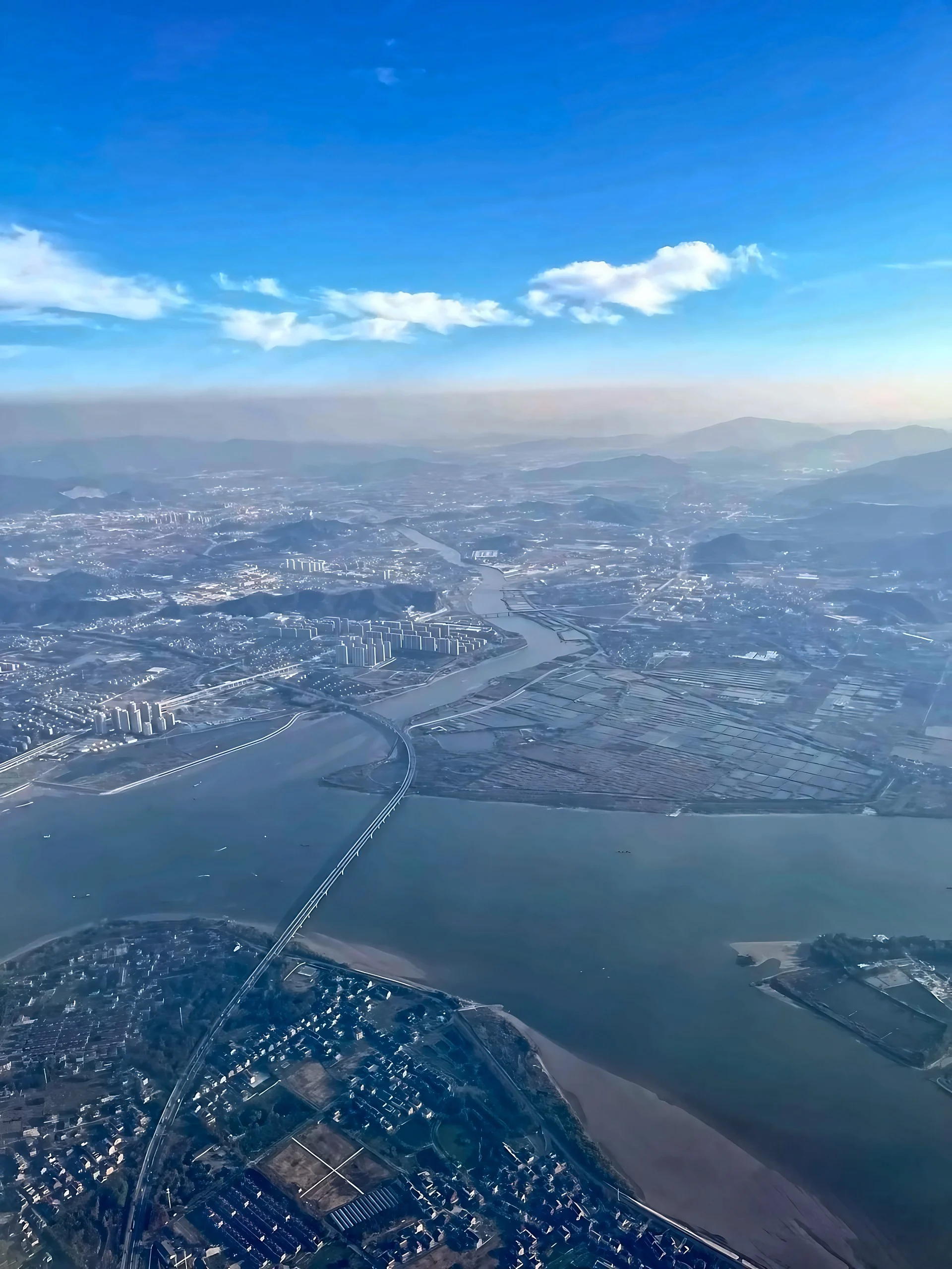 A broad river and long bridge stretch through Hangzhou's riverside settlements beneath blue sky and white clouds, with layered hills in the distance.