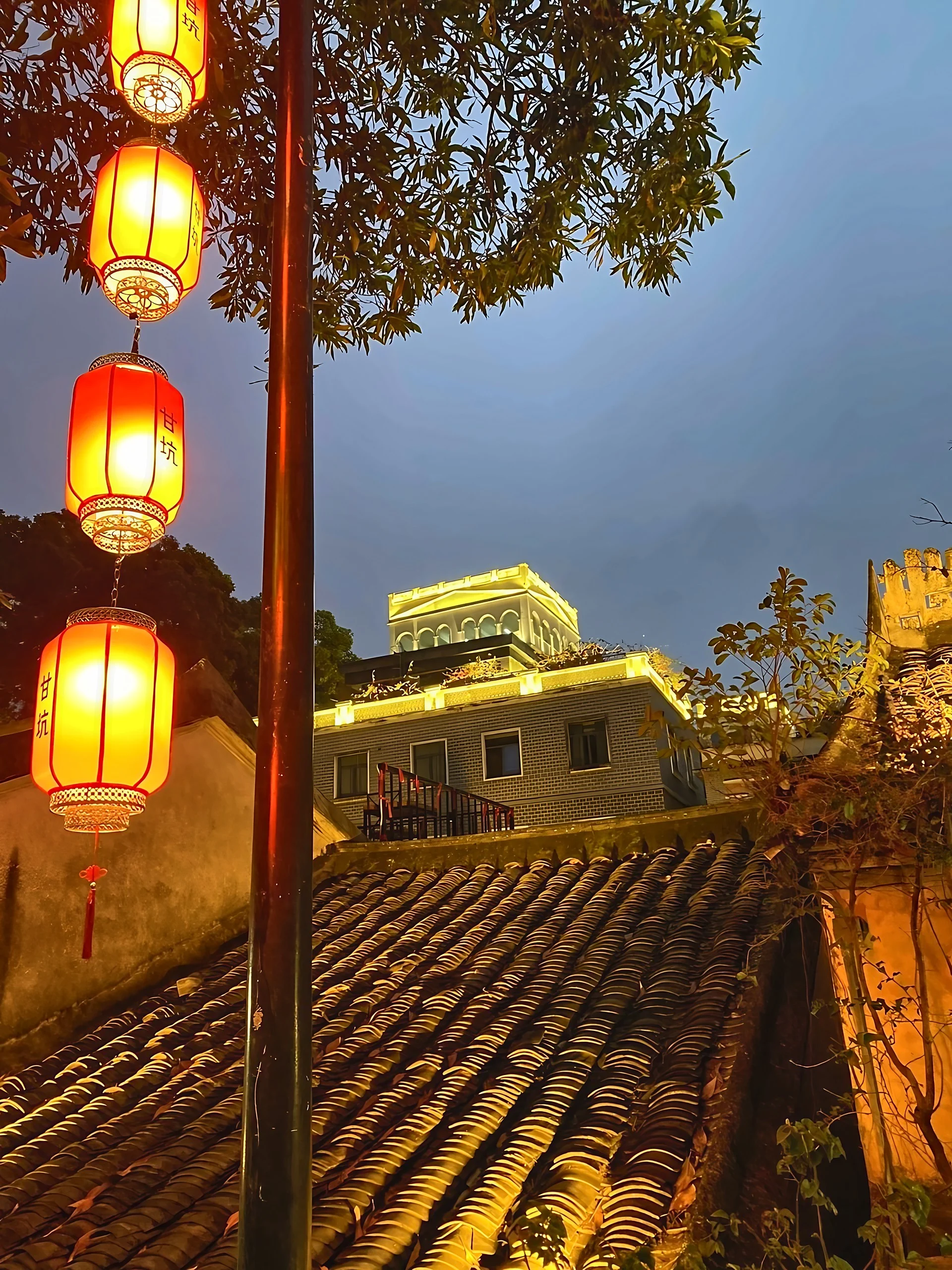 Red lanterns marked Gankeng hang before layered tile roofs and a brick building outlined with golden lights beneath an overhanging tree canopy.