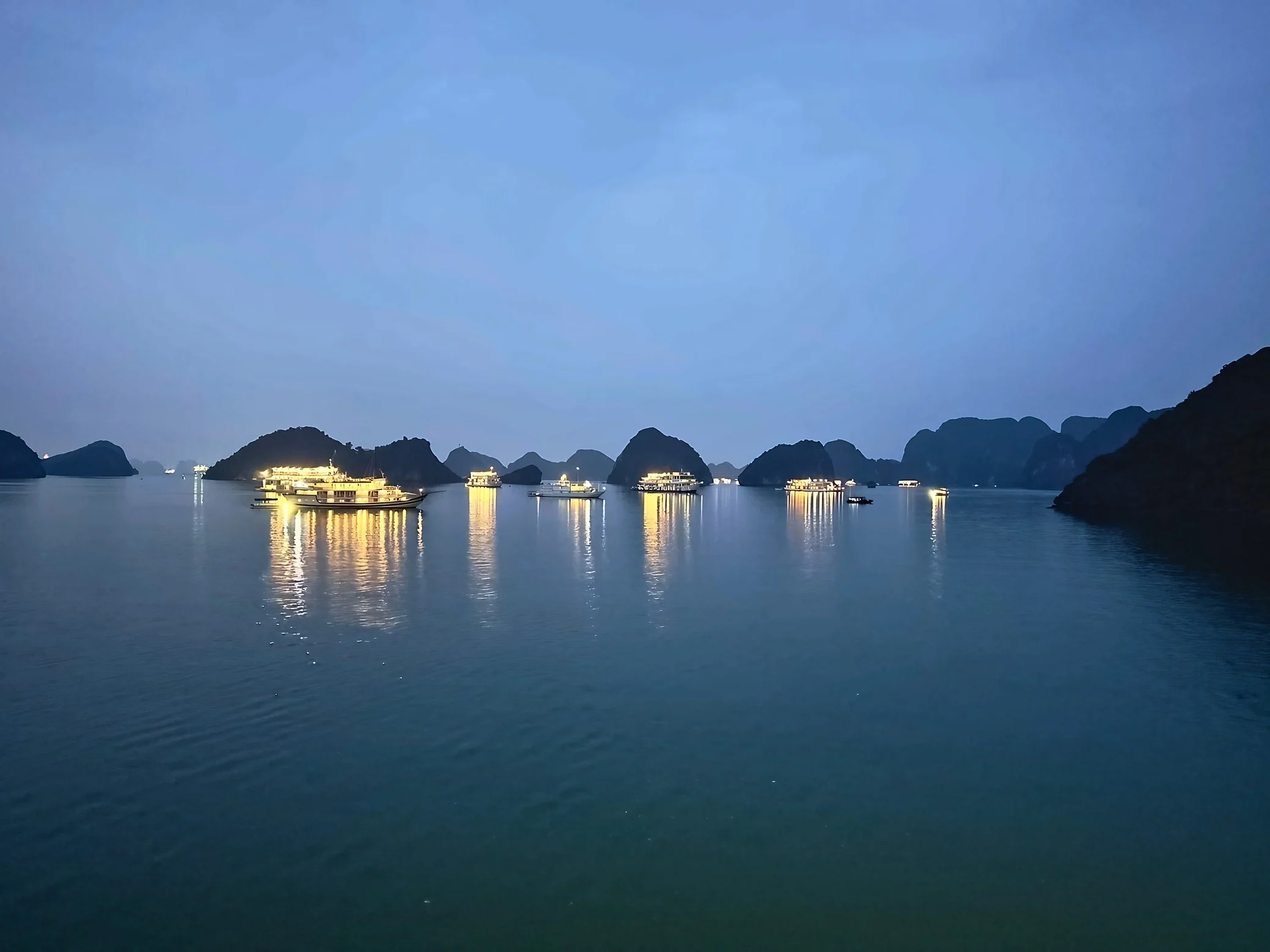 Lit cruise boats and slender golden reflections dot a calm bay surrounded by layered limestone islands.