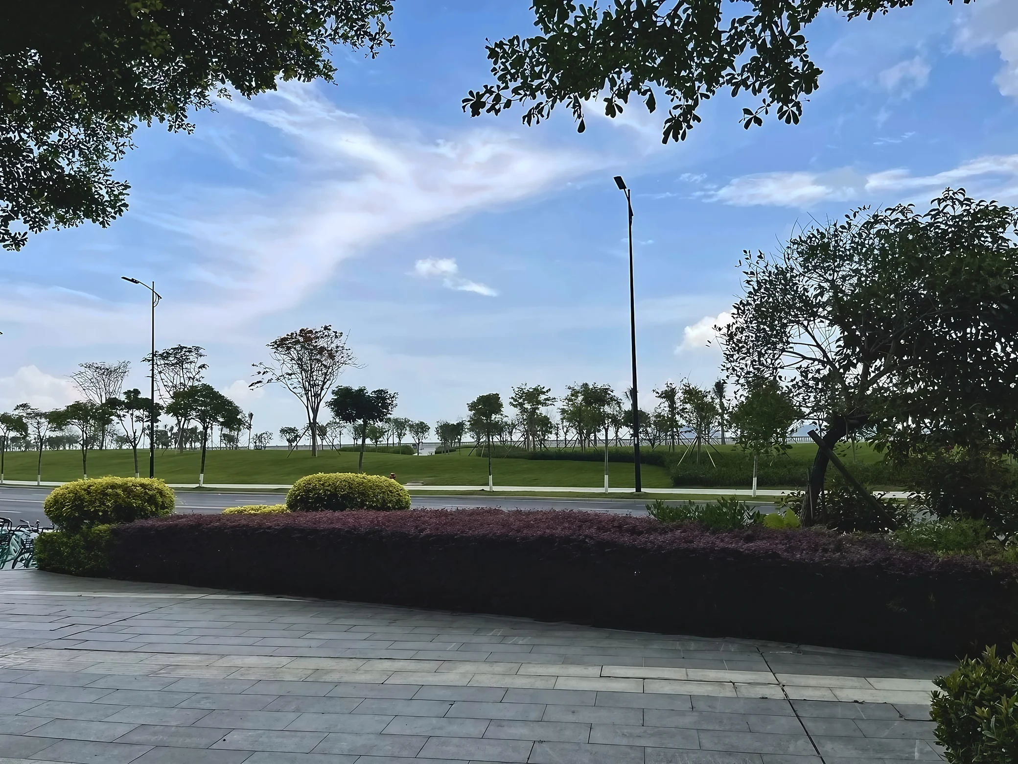 A shaded stone walkway overlooks roadside shrubs, grassy slopes, trees, tall streetlights, and a distant strip of water beneath a blue sky.