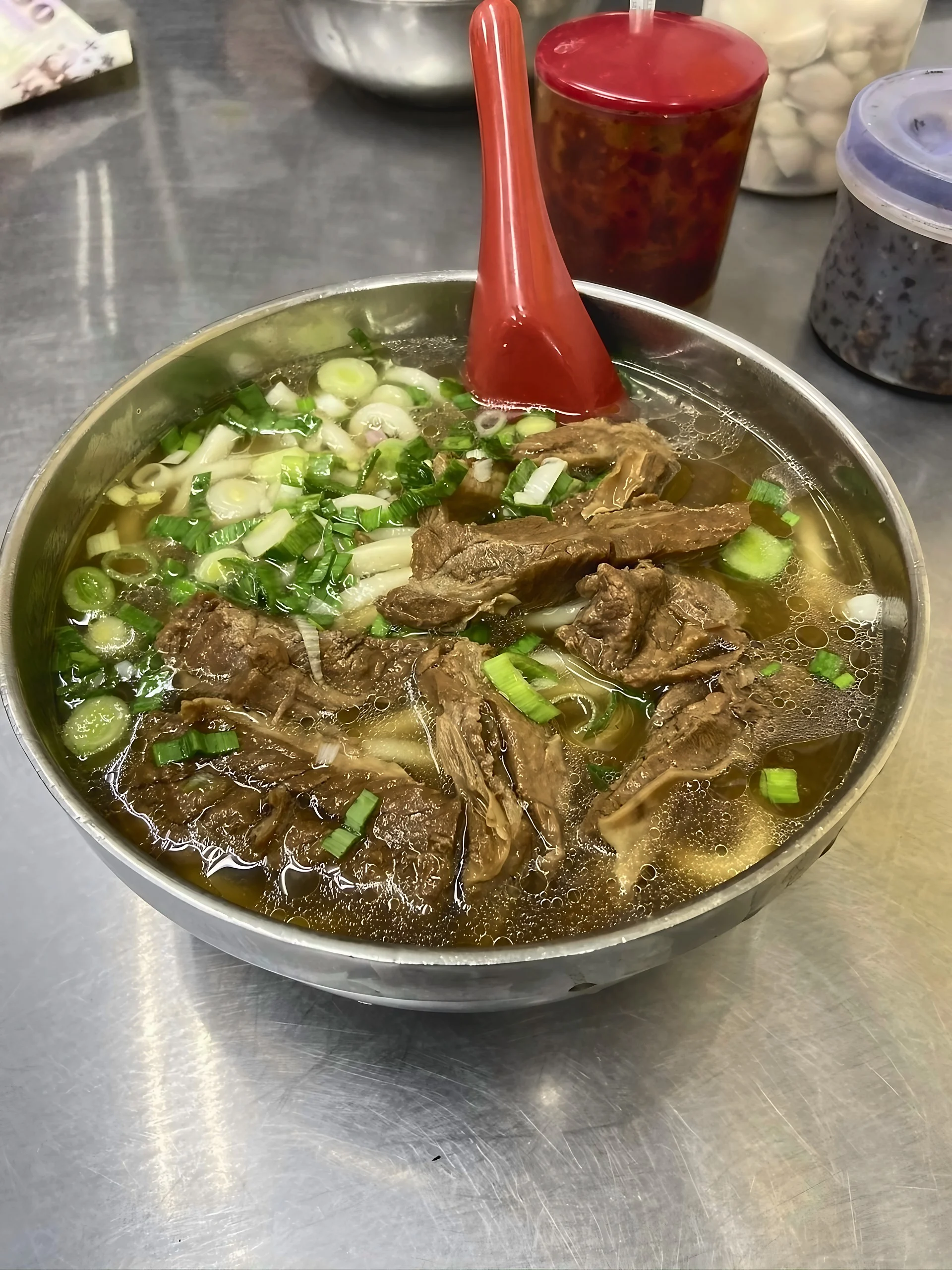A stainless steel bowl of beef noodle soup topped with beef chunks and scallions sits with a red spoon and condiments behind it.