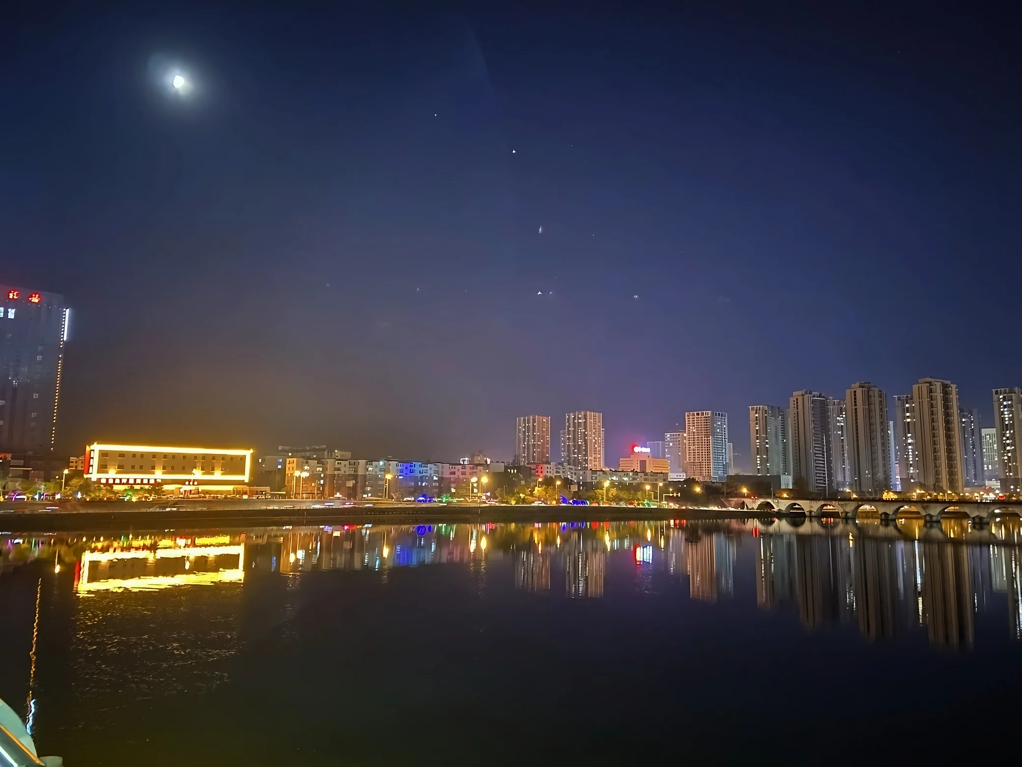 Illuminated riverside buildings and an arched bridge reflect in calm water beneath a deep blue sky.