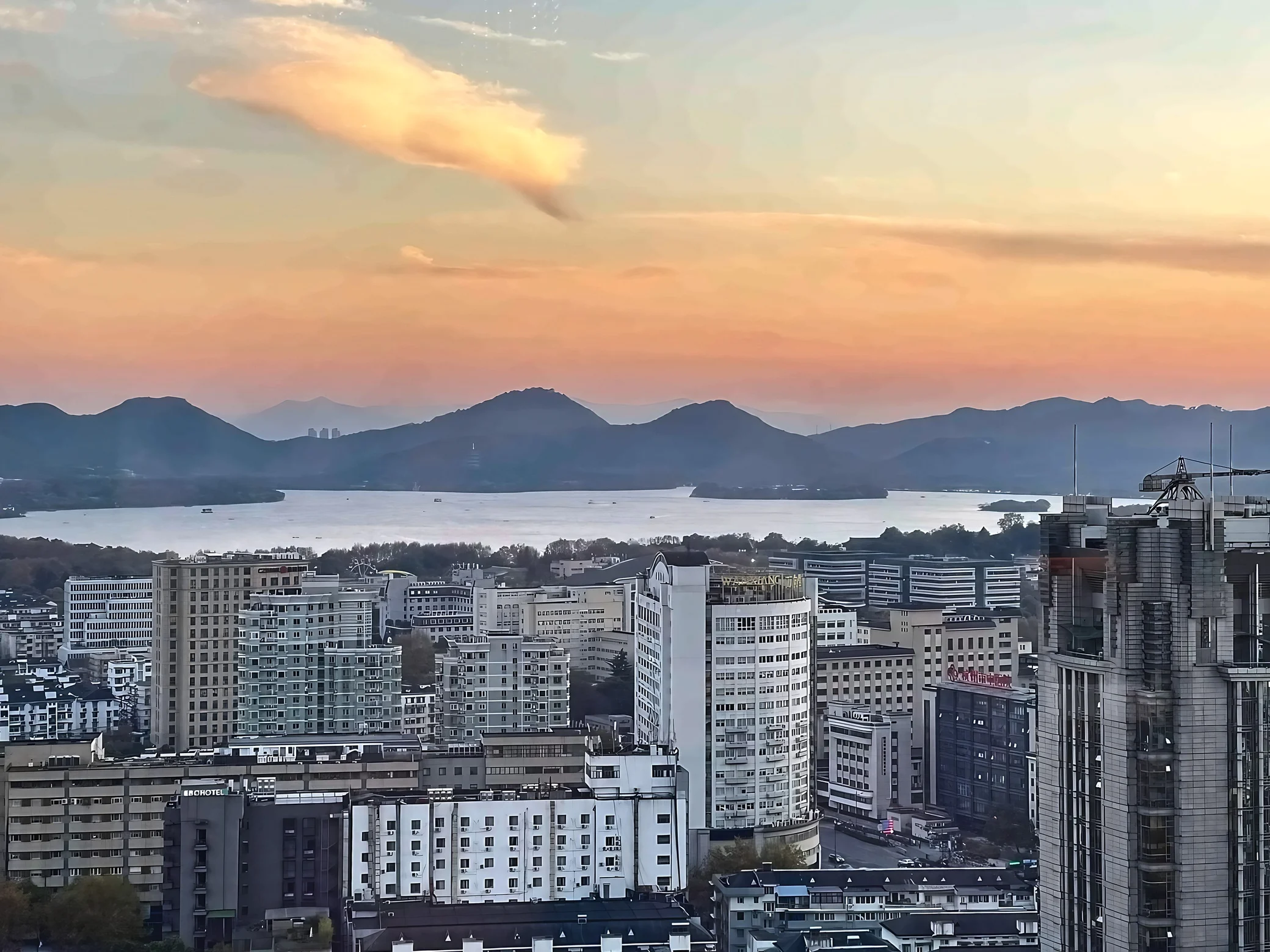 Hangzhou’s buildings stand before West Lake, small boats and layered mountains beneath orange-pink clouds.