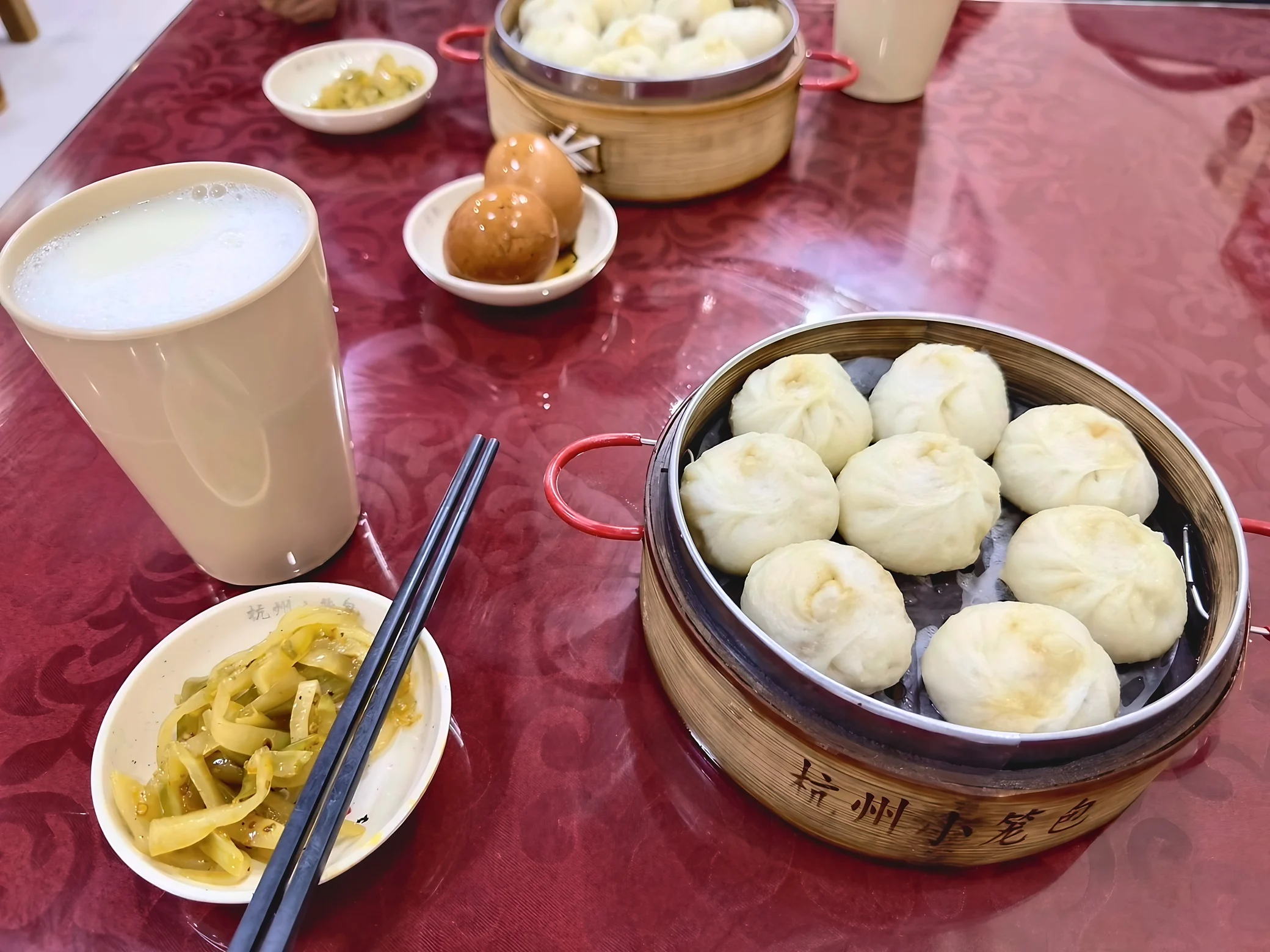 Two baskets of steamed buns, a cup of soy milk, two tea eggs, and small dishes of pickled vegetables sit on a red tablecloth.