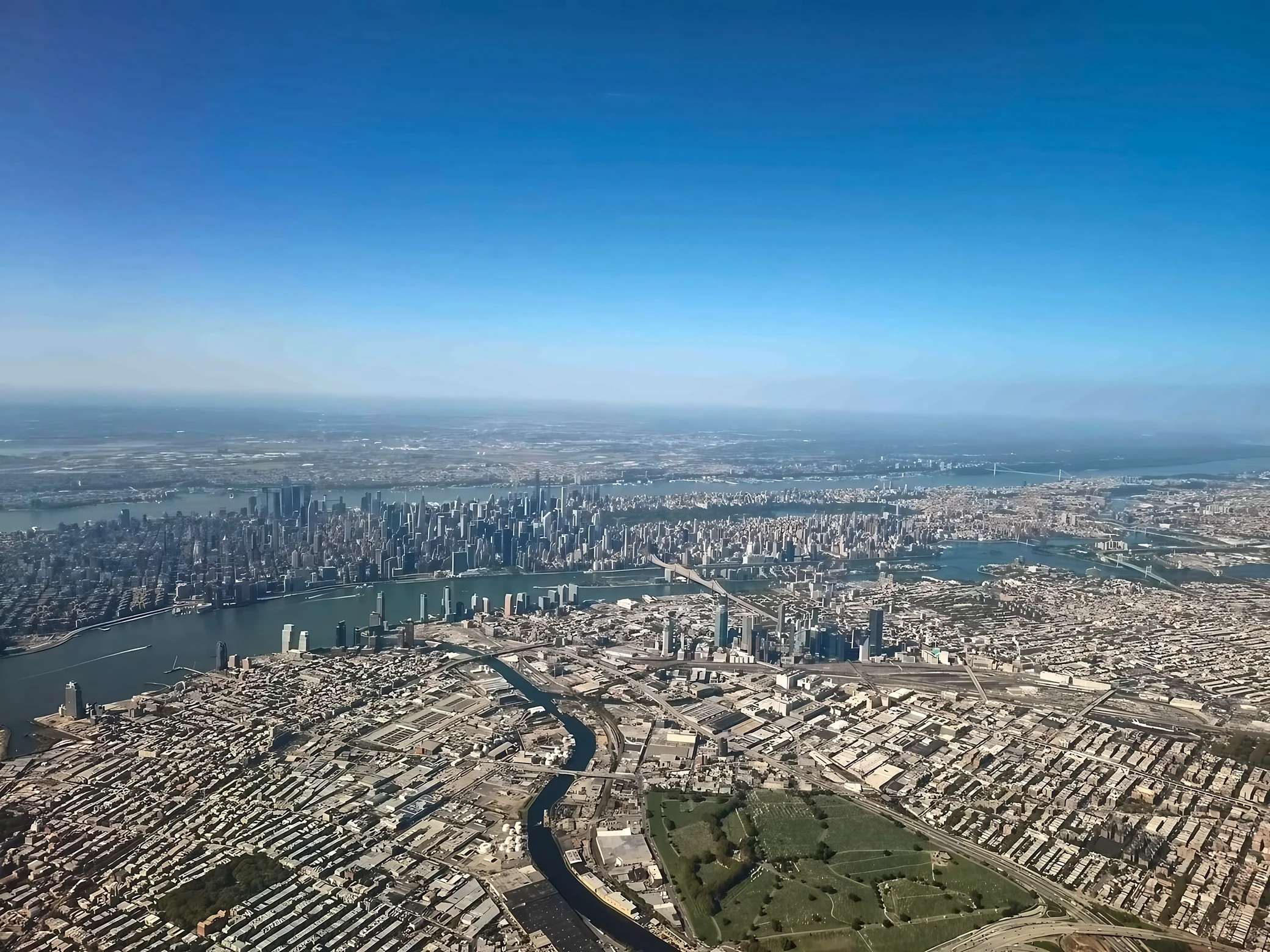Manhattan's skyline rises beyond the East River, with bridges, Newtown Creek, industrial blocks and a cemetery in the foreground.