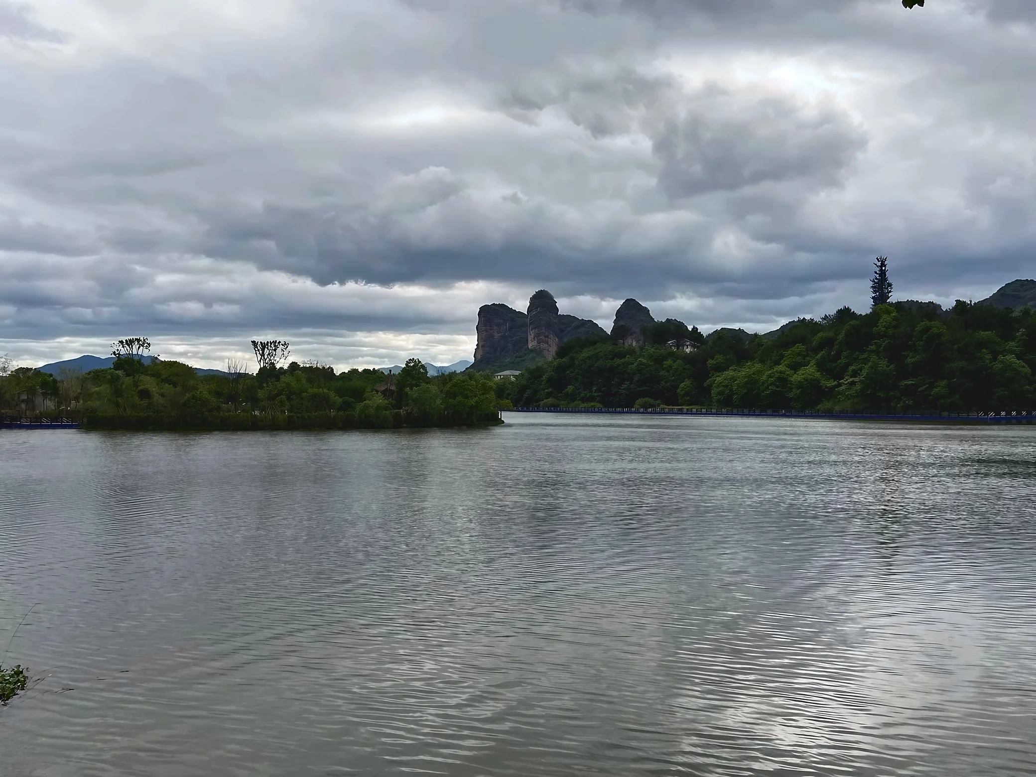 Three prominent Danxia rock peaks rise behind dense woodland and a rippling lake beneath a broad canopy of gray clouds.